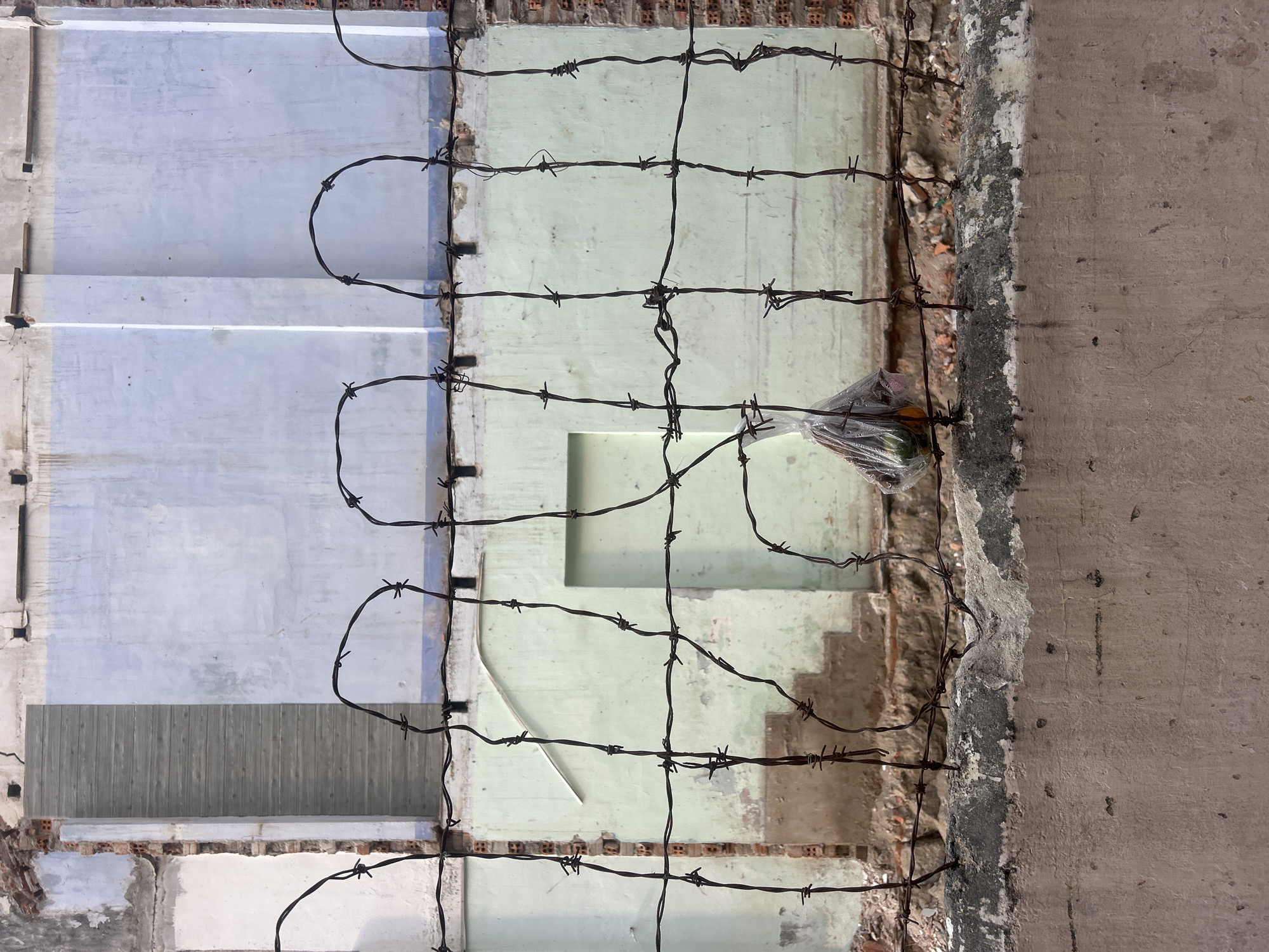 The image depicts a dilapidated building in Nha Trang, Vietnam, partially obscured by a makeshift barbed wire fence. The building's facade is a patchwork of pale blue and green sections, revealing brickwork and signs of decay. The barbed wire is strung haphazardly, suggesting an attempt to secure the derelict structure. A plastic bag containing indeterminate objects is snagged on the wire. The frame is asymmetrical, showcasing the building's deterioration and the improvised security measure.