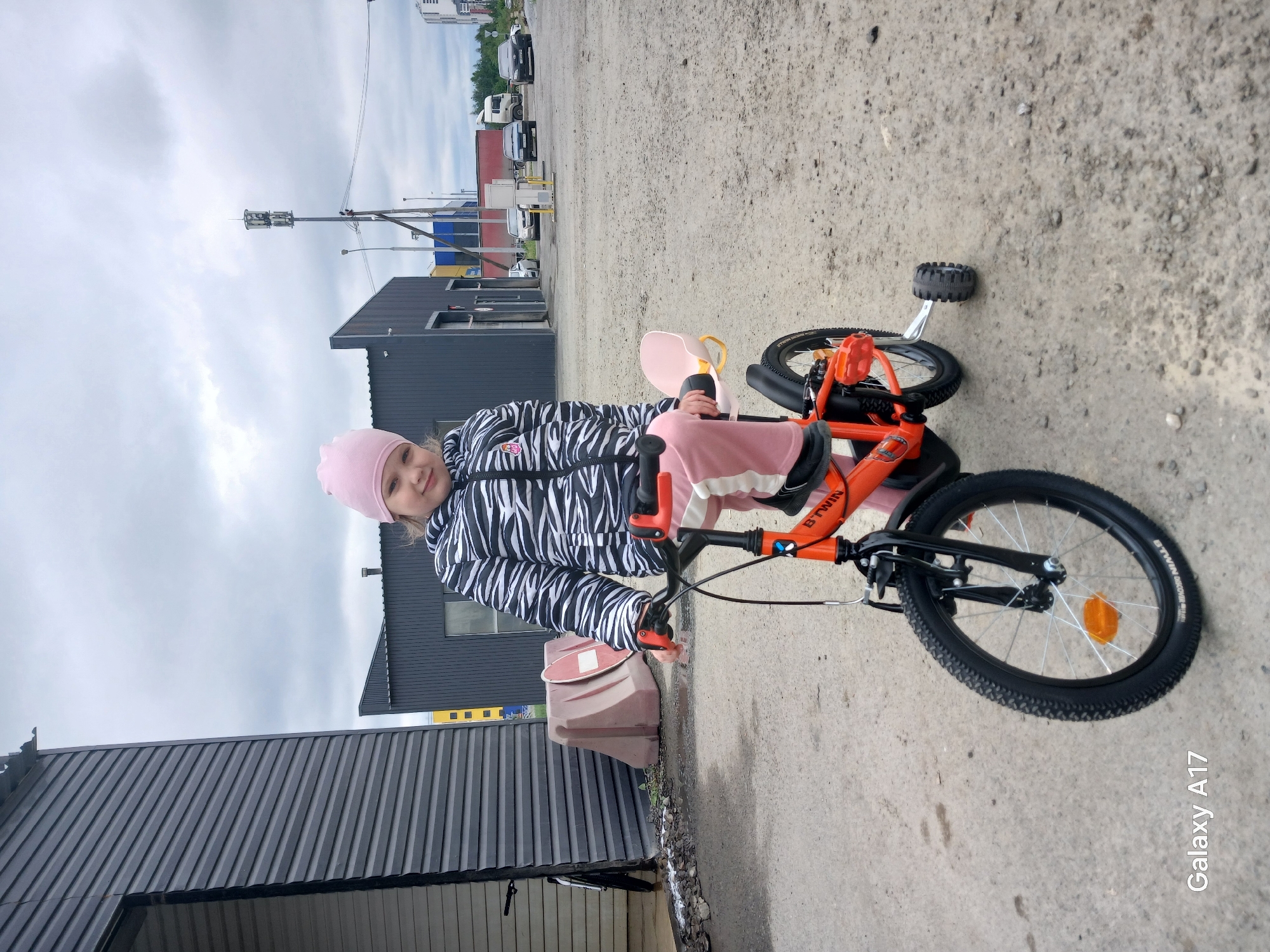 A young girl wearing a pink hat, a zebra-striped jacket, and pink pants is sitting on an orange bicycle with training wheels. The bicycle is on a concrete surface, and the girl is looking towards the camera. Behind her are low industrial-style buildings with dark gray corrugated metal walls. In the distance, there are more buildings and vehicles, obscured by the overcast sky. The image appears to be taken near industrial buildings.