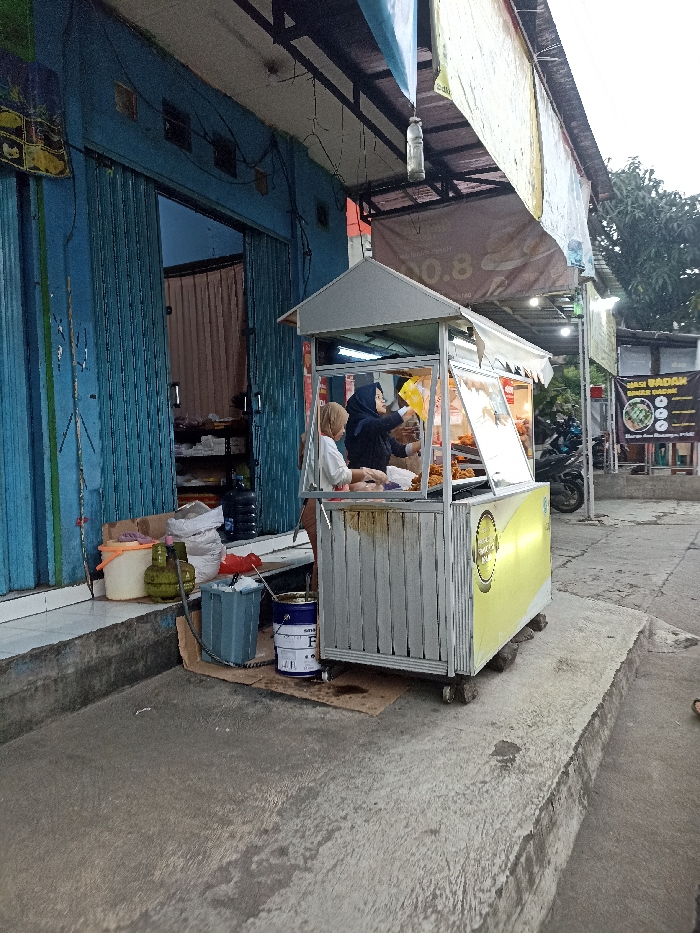 The image depicts a food stall located at a street corner in Jatinangor, Indonesia (latitude: -6.935, longitude: 107.773). Two women stand in front of the stall. One woman, wearing a black headscarf, is handing something to the other woman, who is wearing a white headscarf. The stall is made of metal and has a white metal roof. The stall has a glass display case and shelves. The women are standing in front of the stall, while a motorcycle is parked behind the stall. There are various objects surrounding the stall, including a bucket, a water jug, and a blue paint can. There are also a few bags on the ground near the stall. The stall is located on a sidewalk that is next to a street. The street is paved with concrete. There are trees in the background. The sky is overcast.