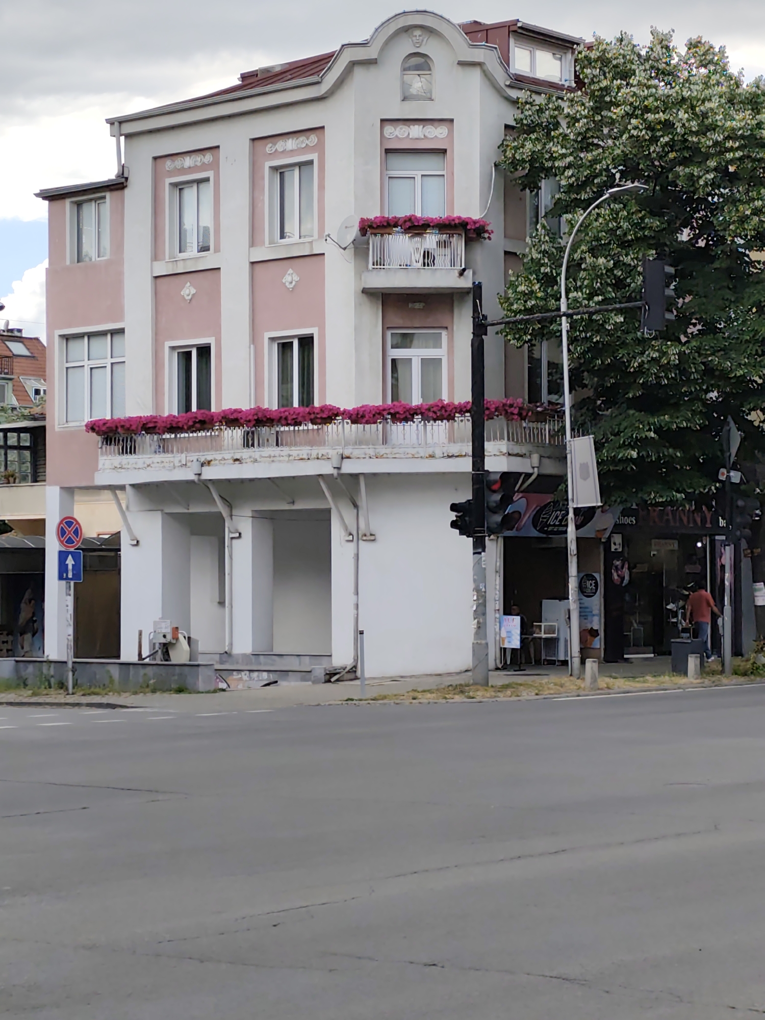 The image shows a multi-story building with pink and white facade, featuring a balcony with pink flowers on the second and third floors. The building is located in Varna, Bulgaria, at the intersection of "Primorski" Boulevard and "Slivnitsa" Boulevard. A traffic light stands in front of the building, next to a large tree. On the ground floor, there is a shop with a sign that reads "ICE" and "KANNY". A person in an orange shirt is walking away from the shop. A traffic sign indicating no stopping and a blue arrow pointing upwards is visible on the left side of the image. The street in the foreground is paved and appears to be a road intersection. The sky is cloudy.