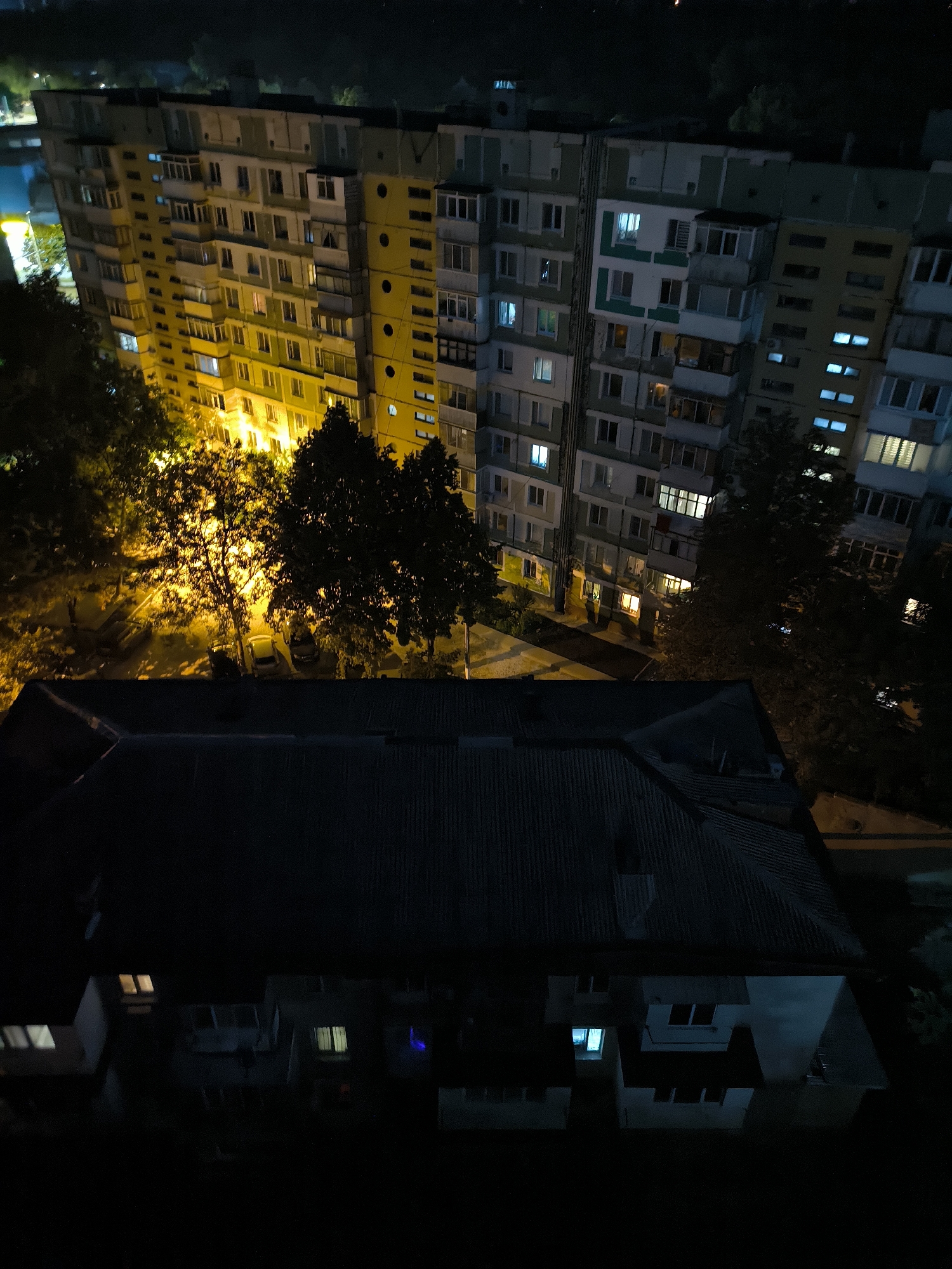 An aerial, night-time view depicts an apartment building in Chișinău, Moldova. The building is illuminated by the interior lights of various apartments, casting a warm glow. Some windows are dark, while others emit light in shades of white, yellow, and blue. Trees with dark foliage are interspersed between the buildings, some of which are also illuminated by streetlights. In the foreground, a darker, smaller building with a corrugated roof is visible. The scene suggests a residential urban environment at night, with the buildings and trees casting shadows and the lights from the apartments creating a sense of occupation and life.