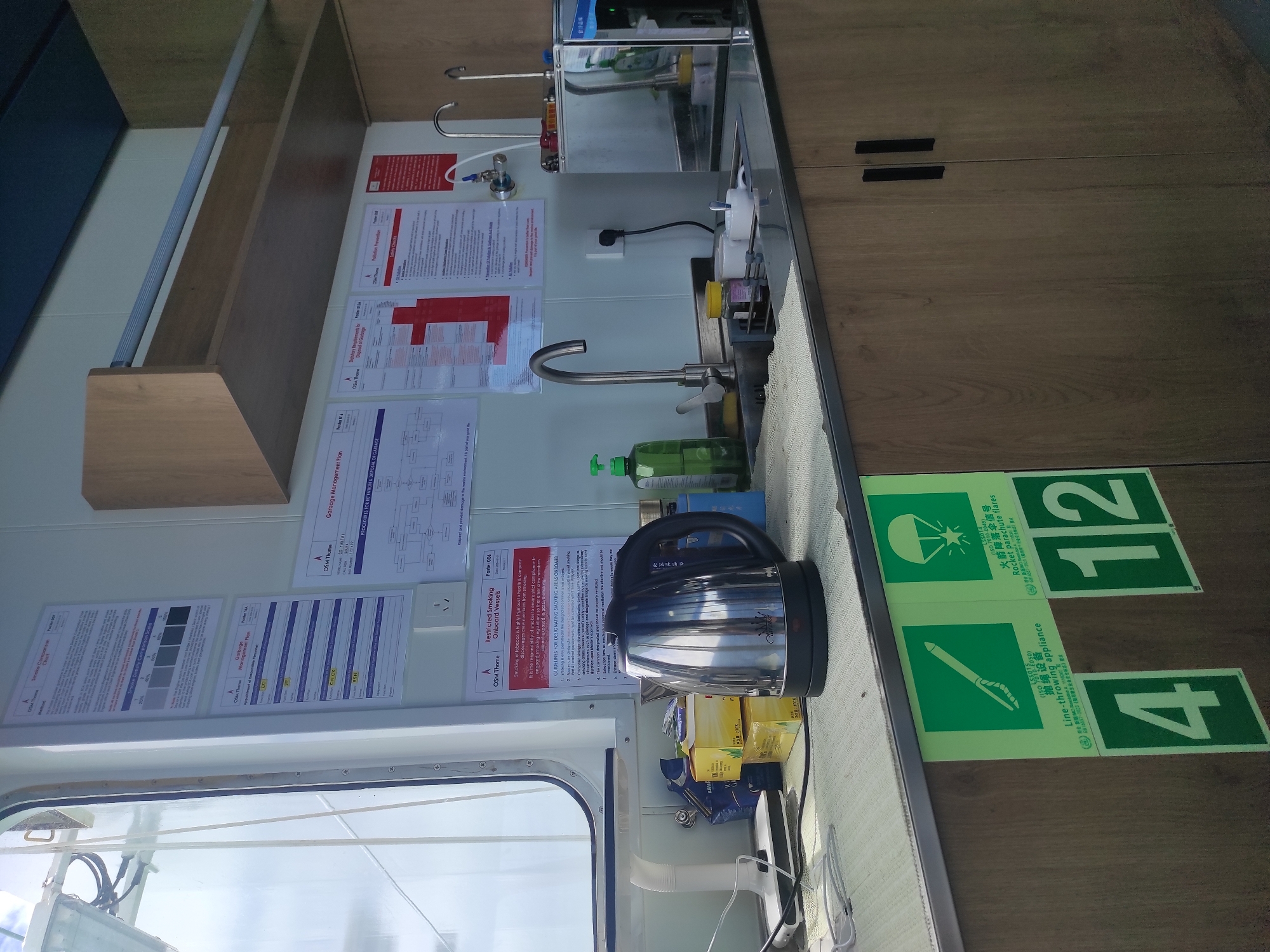 The image depicts a well-lit interior space, possibly a galley or common area on a vessel. In the foreground, a stainless steel electric kettle is prominently placed on a counter next to several boxes of tea. A white electric lamp with a flexible neck is also visible on the counter. To the right of the kettle is a stainless steel sink with a faucet and a green bottle of dish soap. Behind the sink, a water dispenser with a chrome finish is visible.
The wall behind the counter is adorned with several posters. One poster, titled "Restricted Smoking Onboard Vessels," outlines guidelines for designated smoking areas. Other posters are visible, including one with a diagram and text related to "Smoke Density Comparison Scale," and another titled "Garbage Management Plan." Further to the right, a poster on "Pollution Prevention" is partially visible.
The lower portion of the image shows green safety signs on the wooden cabinet doors. These signs include a graphic of a line-throwing appliance and a rocket parachute flare, along with their respective ISO numbers and Chinese text. Large green squares with white numbers "4" and "12" are also affixed to the cabinet doors. The cabinet doors have black handles.
The overall environment appears clean and functional. The lighting suggests an indoor setting with natural light coming from a window on the left side of the frame. The presence of safety posters and equipment suggests a maritime or industrial setting. This scene does not contain any identifiable geographical markers that would specifically place it in Penajam, Indonesia, nor are there any people or specific actions depicted.
