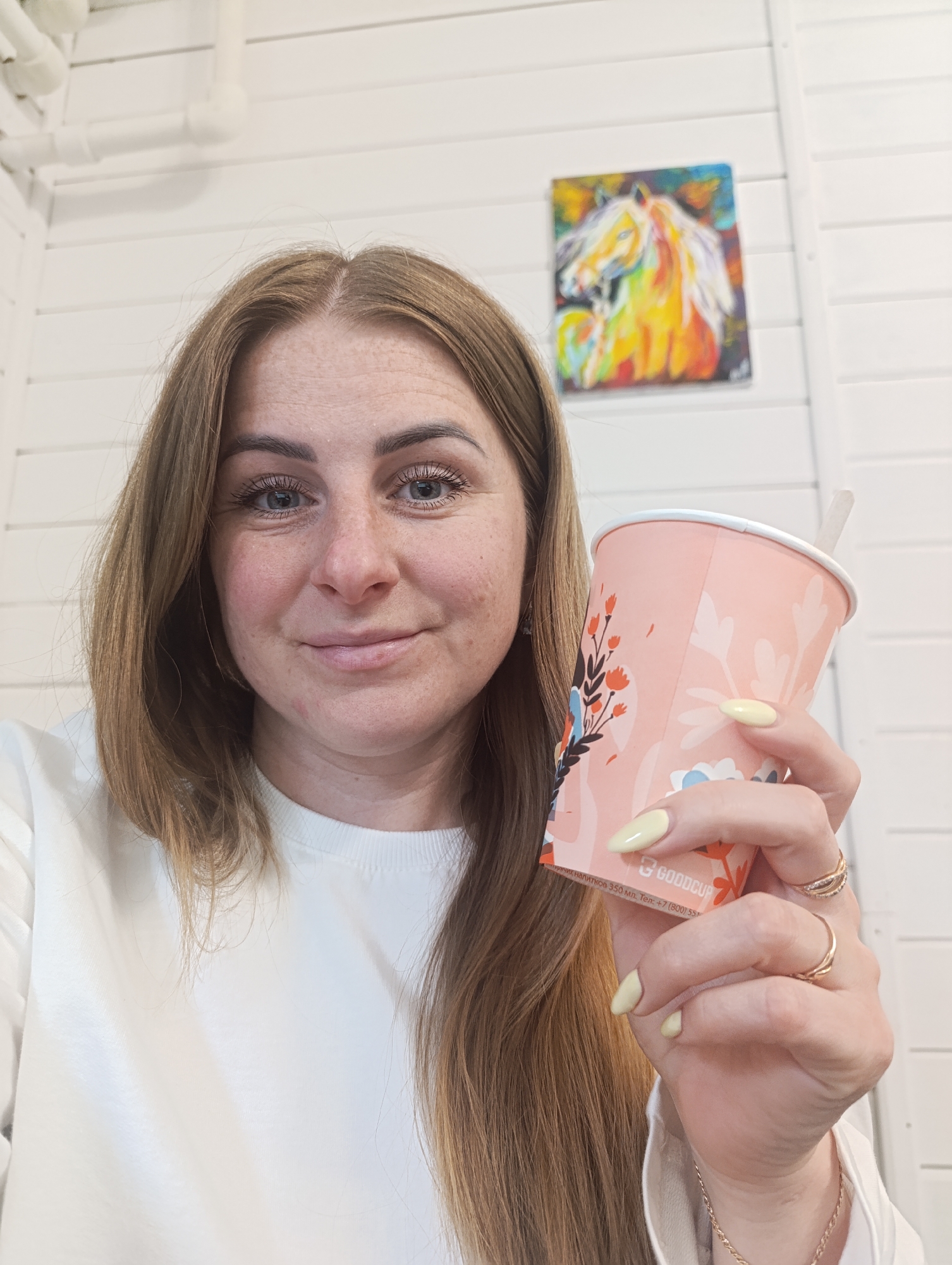 A woman is holding a cup of coffee with a straw in an indoor setting. The woman is facing forward with a slight smile and has shoulder-length brown hair and blue eyes. She is wearing a white sweatshirt. Her nails are painted a pale yellow color, and she is wearing several rings on her fingers. The cup she is holding is pink with a floral design. In the background, there is a white wall with horizontal panels and a painting of a horse in bright colors. There are also some white pipes visible near the ceiling. The location appears to be in the city of Unknown, Unknown.