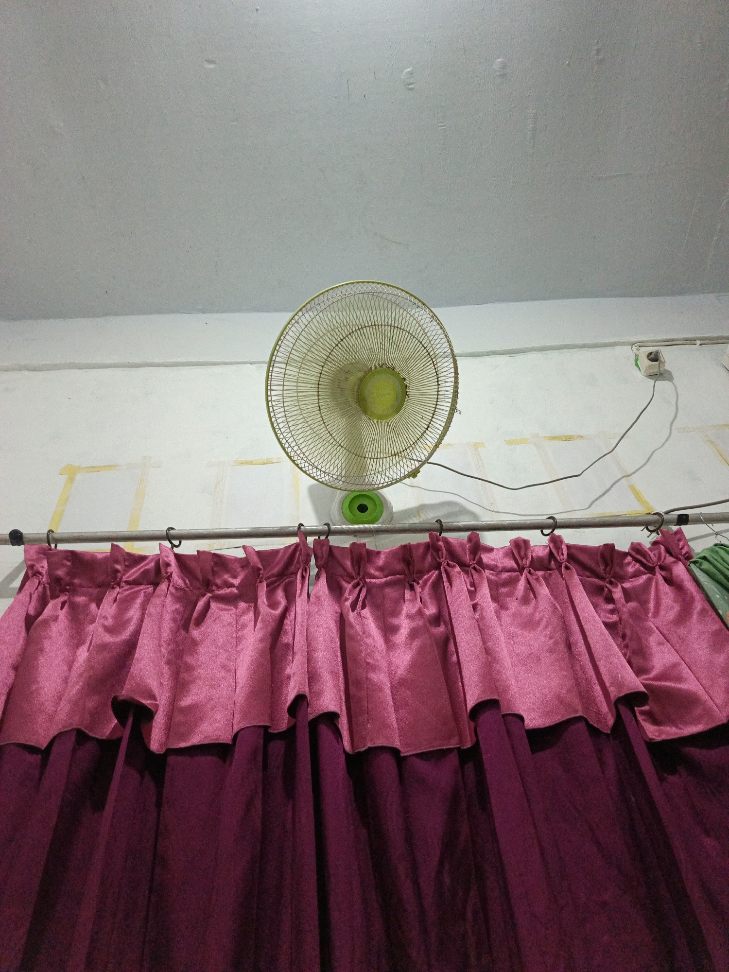 An overhead shot captures a pale green oscillating fan mounted on a white wall above a two-tiered curtain. The top curtain is a lighter, shimmering pink, while the bottom curtain is a deeper, richer magenta. A metal rod supports the curtains, attached to the wall with black clips. To the right of the fan, a white electrical outlet is visible with a black cord extending from it and looping across the wall. The wall itself appears to have been patched in several places with yellow tape, and the ceiling above is a stark white. The scene is indoors, likely a room, with no indication of the specific location within Kecamatan Praya Barat, Indonesia, beyond the provided context.