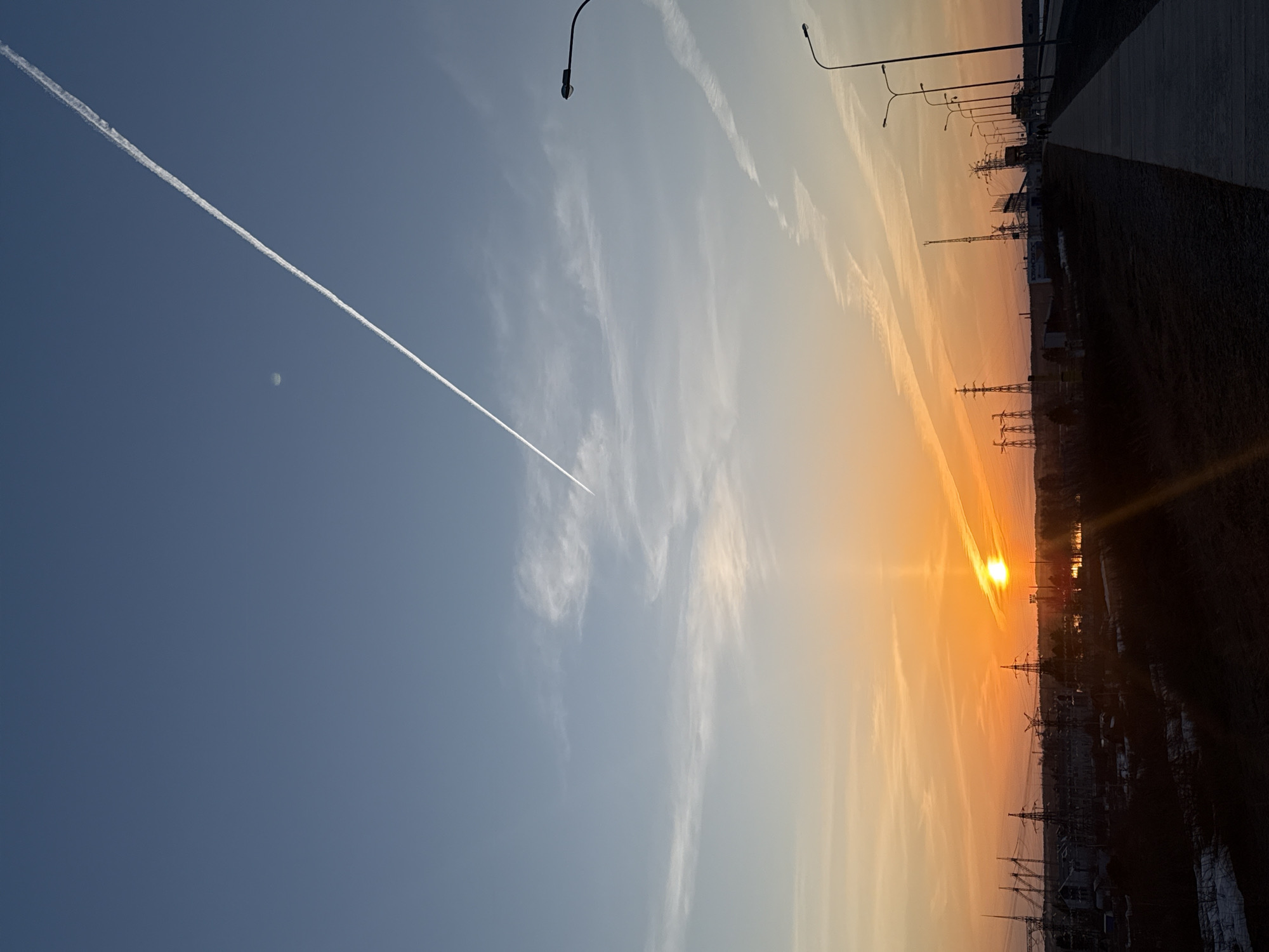 The image captures a sunset over Chaikovsky, Russia, likely taken from an elevated position. The sky is divided into two distinct sections: the left side is a deep blue transitioning to a lighter hue, while the right side is a fiery orange and yellow gradient. A jet contrail cuts diagonally across the left sky, with a small white object (possibly the moon) visible near it.
Below the horizon line are power lines and industrial infrastructure silhouetted against the setting sun. Streetlights are visible along a road or pathway. The lower foreground is dark and indistinct, possibly a field or undeveloped area. The photograph appears to have been taken in a northerly direction, as the sun is setting over the western skyline of the city.