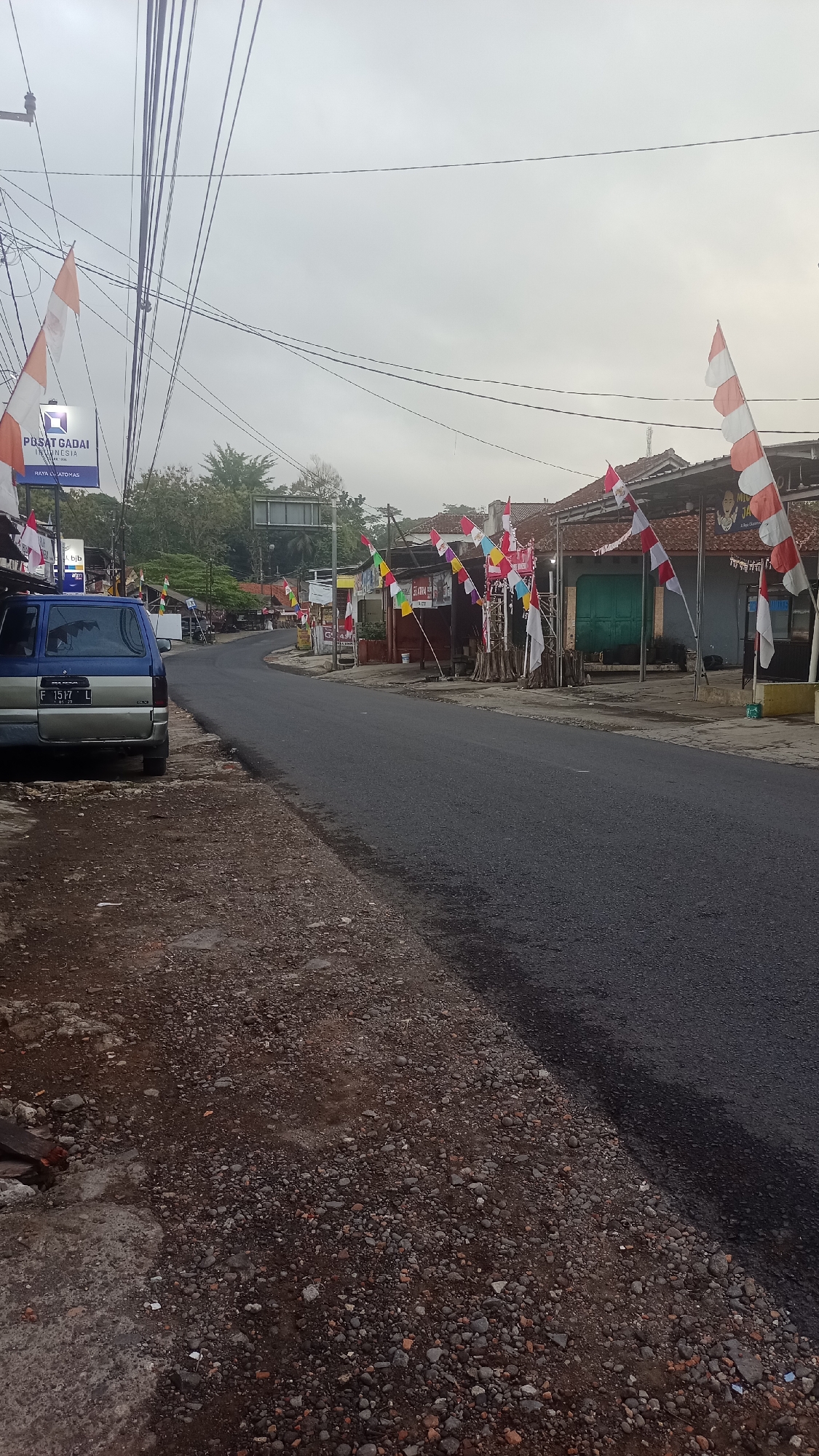 The image depicts a street scene in Pakemitan, Indonesia, decorated with numerous Indonesian flags, indicating a national celebration or event. The road is paved and curves to the left in the distance. On the left side of the road, a blue and silver van is parked, and a sidewalk made of gravel and concrete is visible. A sign for "PUSAT GADAI" (Pawnshop Center) is present. On the right side of the road, several buildings with tiled roofs are lined up. These buildings appear to be shops or small businesses. A green door is visible on one of the buildings. Various Indonesian flags, both large and small, are displayed along the roadside and on poles in front of the buildings. The sky is overcast, suggesting a cloudy day. The overall impression is a quiet street with festive decorations.