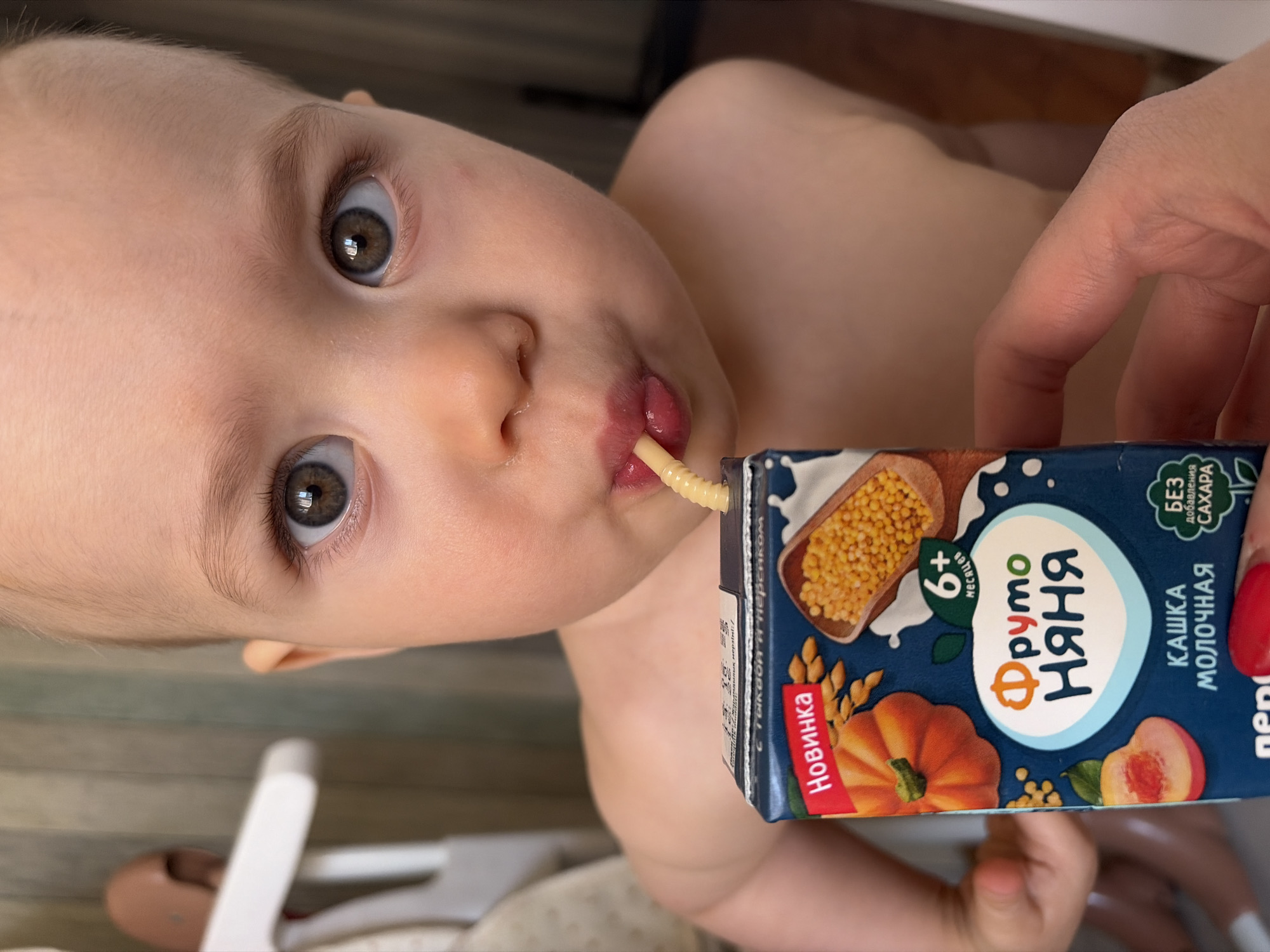 A close-up, medium shot depicts a baby drinking from a box of Fruto Nyanya baby milk cereal in an indoor setting. The baby, with fair skin, brown eyes, and light brown hair, is centered and looking upwards with a curious expression. A straw is in their mouth, connected to the drink box. The hand of an adult is partially visible holding the box of Fruto Nyanya. The box is dark blue with illustrations of millet, pumpkin, and peach, along with Russian text and the brand logo. The background is somewhat blurred, showing elements of what appears to be a high chair and a patterned blanket. The location is Chaikovsky, Russia, inferred from the Russian brand name on the packaging.