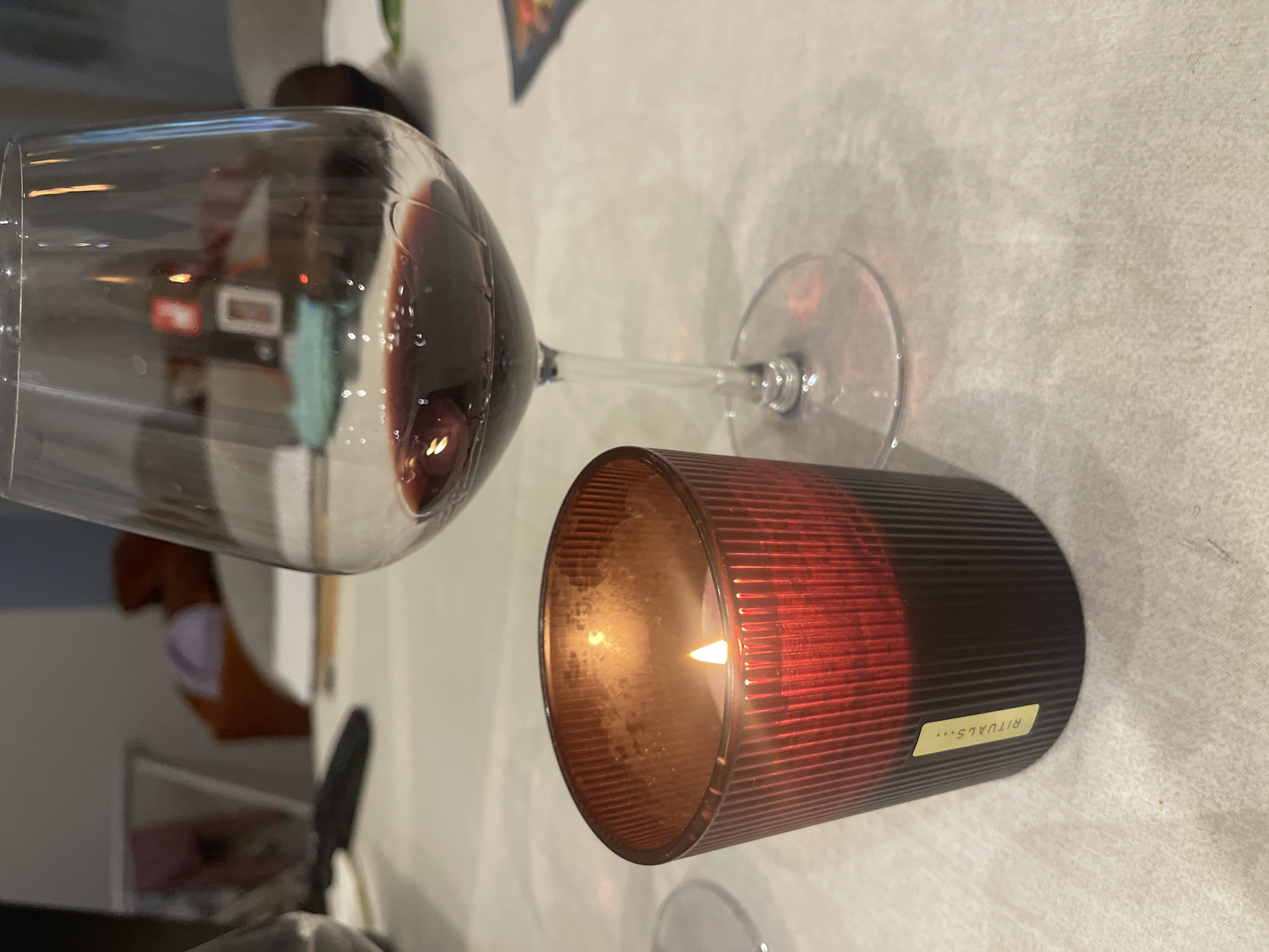 The image depicts a partially filled wine glass and a lit candle on a white surface. The wine glass contains red wine. The candle is in a dark red and black container with vertical ridges, and a small label that reads "RITUALS..." is visible. The location is Unknown, Unknown. No people are directly visible in the frame, but the reflection in the wine glass suggests a domestic setting. The environment is dimly lit.
