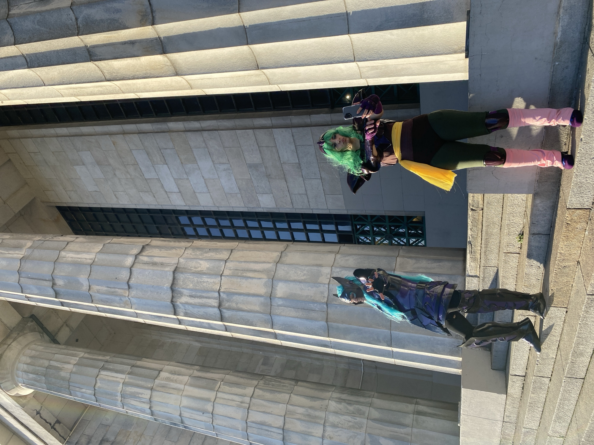 Two people in cosplay costumes stand on a ledge of a building with classical architecture. One is wearing a green wig, purple and brown costume with a yellow sash, pink leg warmers and is holding a cell phone. The other person is wearing a blue wig and a black and blue costume. The building has large columns and a grid of windows. The location is in the city of Buenos Aires, Argentina.