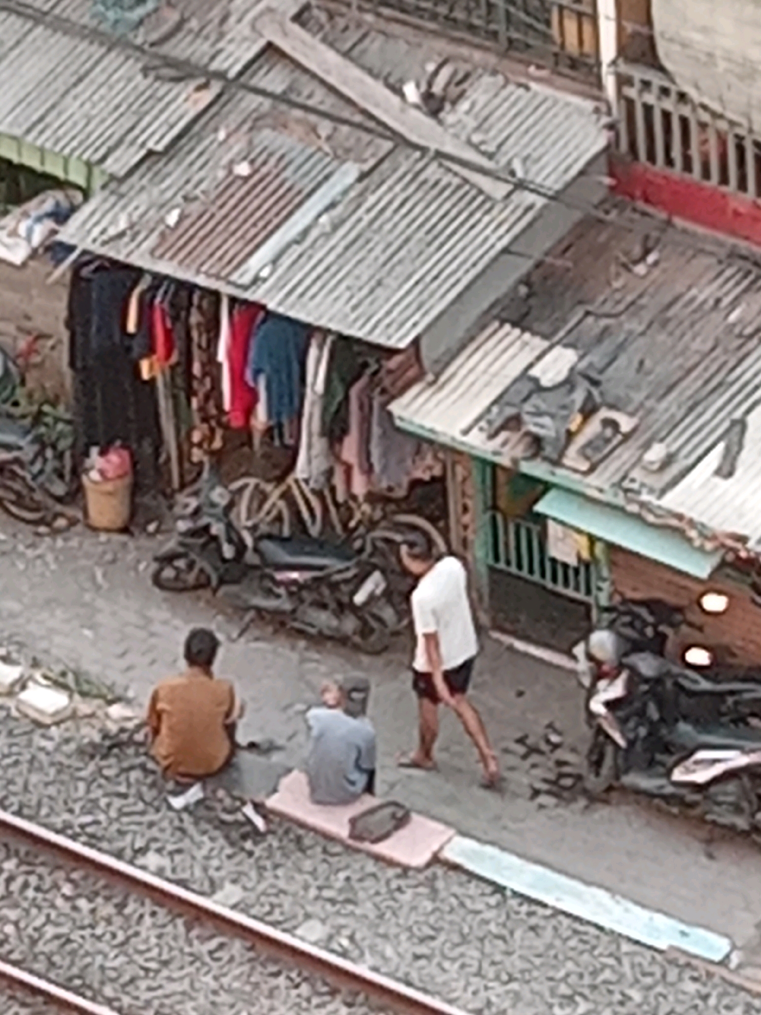 The image depicts a scene in a densely populated urban area, likely a slum, in Surabaya, Indonesia. In the foreground, railroad tracks run horizontally. Adjacent to the tracks, two men are seated on the ground. One man, wearing a brown shirt, is facing away from the camera. The other man, in a grey shirt, is also facing away and appears to be slightly behind the first.
Behind the men is a makeshift structure with a corrugated tin roof, typical of informal settlements. Various items of clothing are hanging on display outside this structure, suggesting it may be a small stall or part of a residence. Several motorcycles are parked around the area, indicating modes of transportation common in the region.
Further back, another man wearing a white t-shirt and shorts is walking away from the camera. He is dressed casually and appears to be moving through the settlement. The overall environment is characterized by clutter, makeshift construction, and a sense of crowded living conditions. The lighting suggests it is daytime.