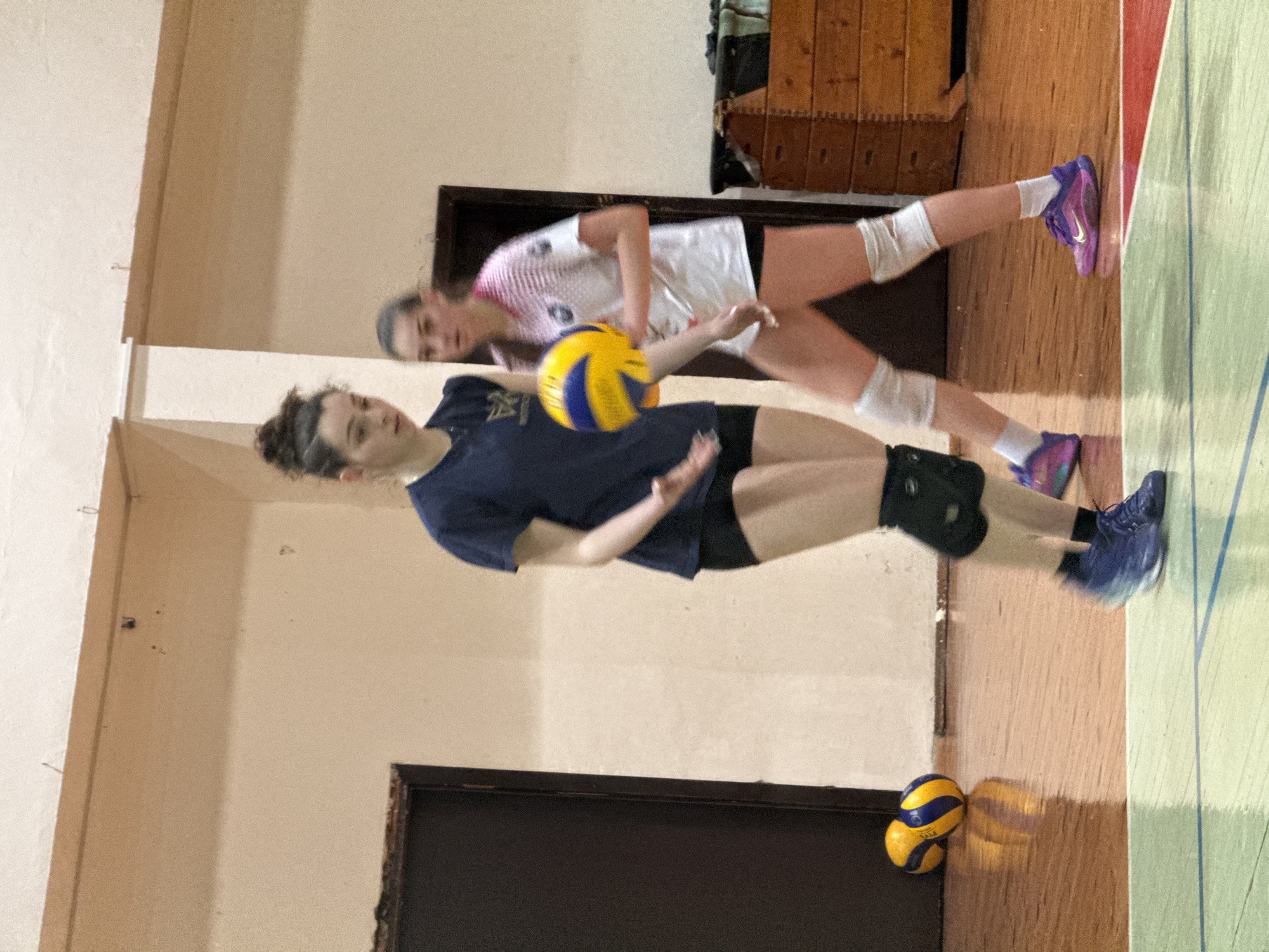 Two female volleyball players are practicing indoors. One player, wearing a dark blue t-shirt and black shorts with a knee pad on one leg, is hitting a yellow and blue volleyball. Another player, wearing a white and pink jersey with white knee pads, stands behind her. Two additional volleyballs lie on the floor near a door. The location appears to be a gymnasium, with a wooden floor and a wooden cabinet in the background. The location is unknown and cannot be determined based on the image.