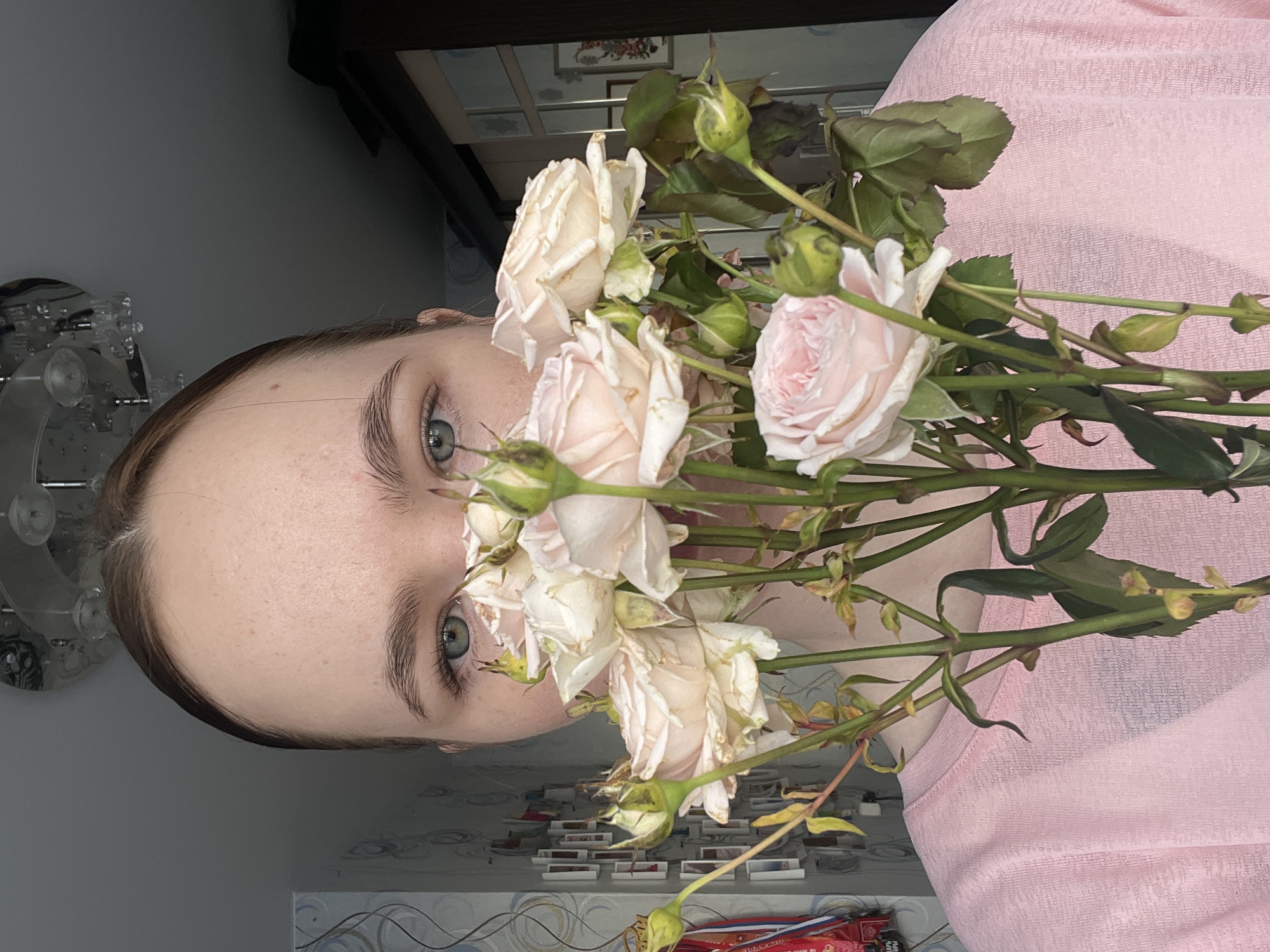 A fair-skinned individual with light-colored eyes is positioned behind a bouquet of pale pink and white roses. The individual is wearing a pink top. The background includes a partially visible wall, ceiling light fixture, and a cabinet. It is difficult to definitively confirm the location as Chaikovsky, Russia without additional visual or contextual information from the image.