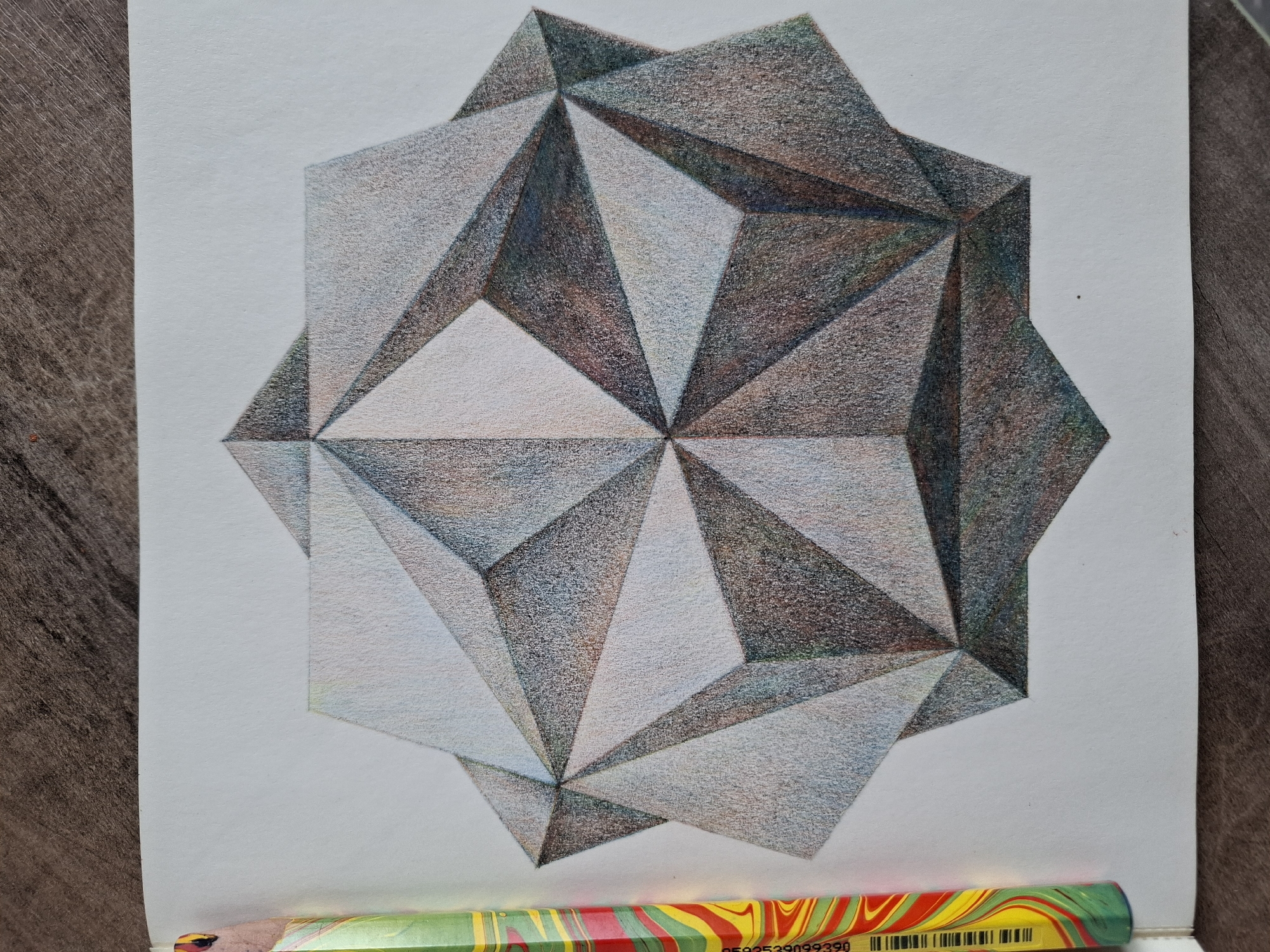 A colored pencil drawing of a complex geometric shape is presented on white paper. The shape appears to be a star or a polyhedron with many facets. The drawing utilizes shading to create a sense of three-dimensionality, with some faces appearing lighter and others darker, suggesting light source from the top left. The colors used are predominantly grays, blacks, and whites, with subtle hints of blue and brown in the shading. A brightly colored pencil, with a marbled pattern of red, yellow, green, and white, lies to the left of the drawing, partially in frame. The drawing is placed on a wooden surface. There is no identifiable location in the city of Tyumen, Russia, depicted in this image.