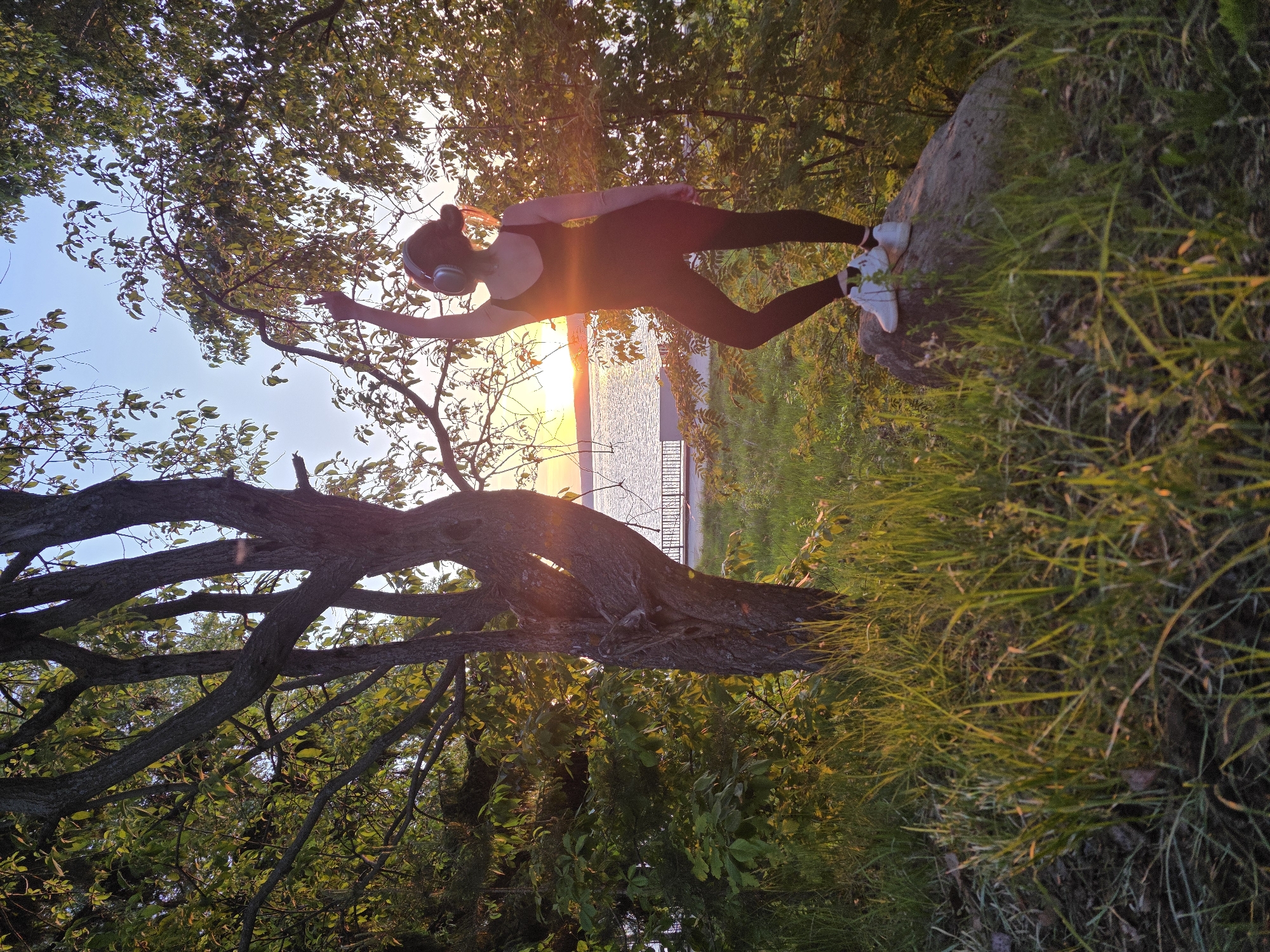 A woman wearing a dark-colored full-body suit and white sneakers stands on a large rock near a lake. She has headphones on and one arm outstretched towards a tree branch. The setting sun is visible behind the lake. The location is Chaikovsky, Russia.