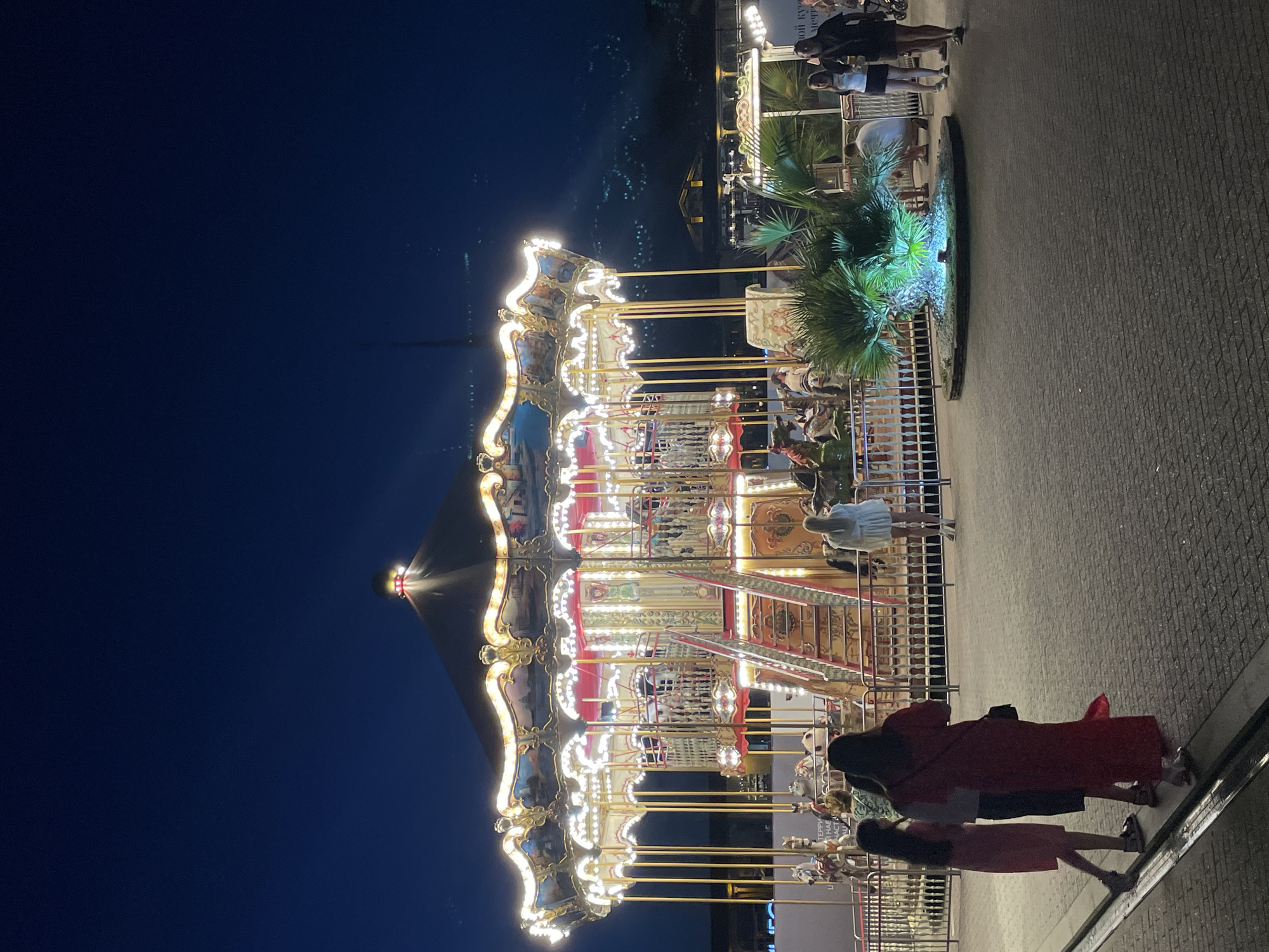 This is a nighttime, eye-level, outdoor, full-shot photograph of a carousel at an amusement park, likely in Sochi, Russia, due to the presence of the illuminated mountains in the background, which are characteristic of the city's landscape.
The carousel, a central feature of the image, is brightly lit with numerous golden lights and adorned with intricate painted panels depicting pastoral scenes. Its multi-tiered structure is supported by ornate golden columns. Several horses, designed for riding, are visible on the carousel's platform. A red staircase leads up to the carousel entrance.
In the foreground, four individuals are walking on a paved area. On the left, two young females, one in a pink dress and the other in a green and white patterned top and dark skirt, are walking away from the camera. To their right, a woman in a long red dress is also walking away from the camera, carrying a black handbag. Further right, a woman in a white dress is standing, facing towards the carousel.
To the right of the carousel, a palm tree is illuminated with a vibrant blue light. Behind it, a group of people are standing and walking, including a man in a white t-shirt and dark shorts and two women in casual attire. In the far background, beyond the amusement park structures, illuminated mountains rise against a deep blue night sky. Buildings with lights are scattered across the mountainside, suggesting a densely populated area. The overall atmosphere is festive and illuminated, with the carousel serving as the main attraction.
