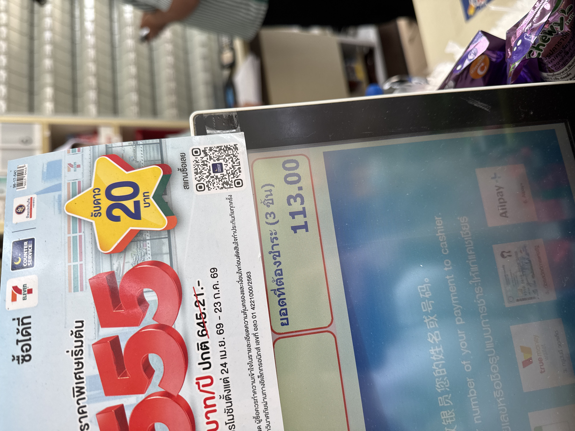 The image depicts a close-up view of a point-of-sale (POS) screen at a 7-Eleven store. The screen displays promotional information and a payment prompt in Thai. Prominently featured are large red numbers "555" and a yellow star graphic with the text "รับดาว 20 บาท" (Receive 20 stars/baht). Below this, text indicates a promotion period from May 24th to June 23rd, 69 (likely 2024). The screen also shows "ยอดที่ต้องชำระ" (Amount to pay) followed by "113.00" in large numbers. Further down, there's a prompt in Chinese and English: "员工您的姓名或号码. number of your payment to cashier." and "เลขหรือชื่อรูปแบบการชำระ. ให้แก่แคชเชียร์." at the bottom, offering payment options like TrueMoney Wallet and Alipay. In the background, shelves with products are visible, and a person wearing a striped shirt is partially visible to the right, holding a mobile phone. The overall scene suggests a transaction taking place at a retail establishment.