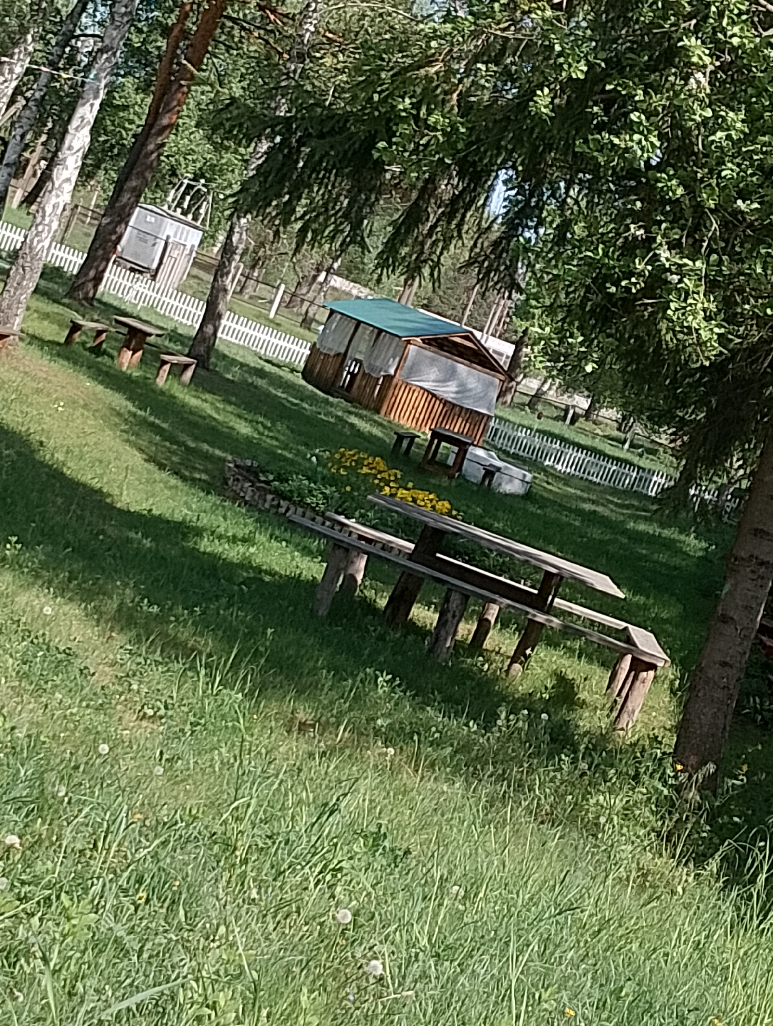 Here is a description of the image:
The image shows a grassy field with picnic tables, trees, and buildings. A white picket fence runs along the background. The scene appears to be in a park or recreational area in the city of Unknown, Unknown.