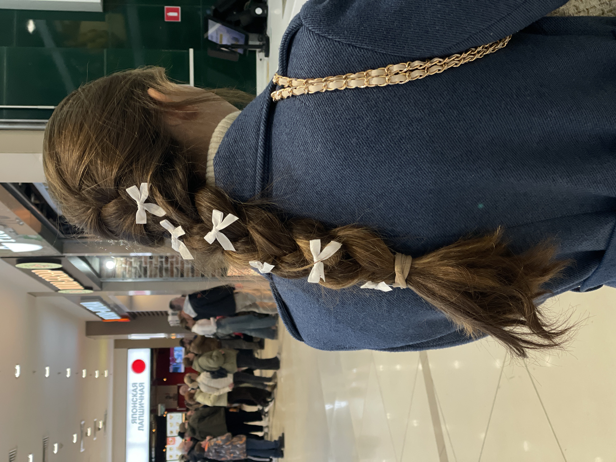 A girl with brown hair is wearing a navy-blue jacket. Her hair is braided with small white ribbons tied along the braid. The jacket has a gold chain detail. The background appears to be a shopping center, with people walking around and a restaurant visible with signage in a foreign language. The location of the city is unknown.