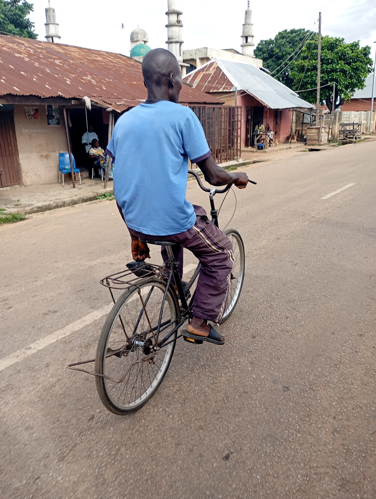 A man rides a bicycle down a paved street in Shendam, Nigeria, with buildings and trees visible in the background. The man, wearing a blue t-shirt and dark pants with stripes, is seen from behind, with his left hand on the handlebar. The bicycle has a metal rack over the rear wheel. In the background, there are several buildings with corrugated metal roofs, some of which appear to be shops or homes. A mosque with minarets is also visible, suggesting a predominantly Muslim community. The street is lined with some vegetation, including a large tree. The sky is overcast.