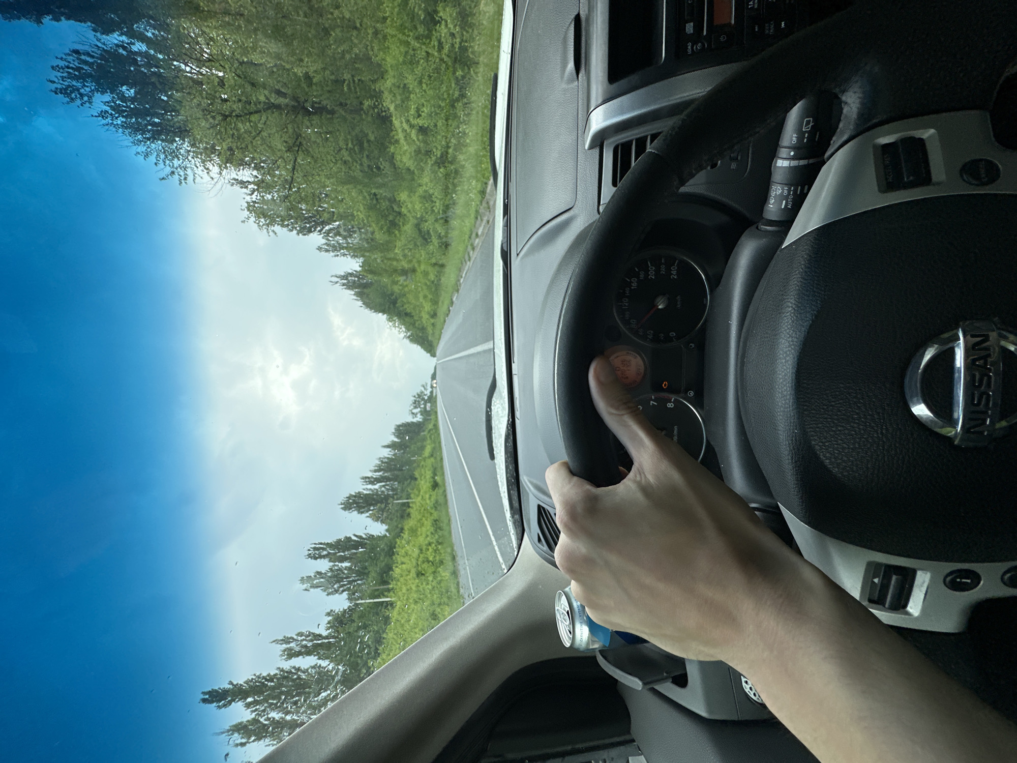 The image shows a first-person perspective from inside a car, likely driving on a highway. A hand is visible on the steering wheel, which has a Nissan logo in the center. The dashboard displays a speedometer with readings up to 240 km/h and other indicators. A windshield wiper control stalk is also visible.
Through the windshield and the side window, a bright, sunny sky with some clouds is seen. Lush green trees line both sides of the road, suggesting a rural or suburban setting. The road appears to be asphalt with lane markings. The overall impression is a daytime drive under pleasant weather conditions.
The visible text includes "NISSAN" on the steering wheel, and various labels on the dashboard controls, such as "ACCEL/RES", "LOAD", and "OFF", "AUTO", indicating the car's features. There is also some text on a small object hanging from the dashboard, possibly a car air freshener, which includes "-CARNITINE VITAMIце".
Given the provided location context of Shatava, Ukraine, this image captures a scene of someone driving through a scenic route in the Ukrainian countryside on a clear day.
---
**Note:** The provided OCR output shows some Ukrainian characters and what appears to be a brand name or product description. The description adheres to the factual and concise requirements, incorporating visible text and scene details.