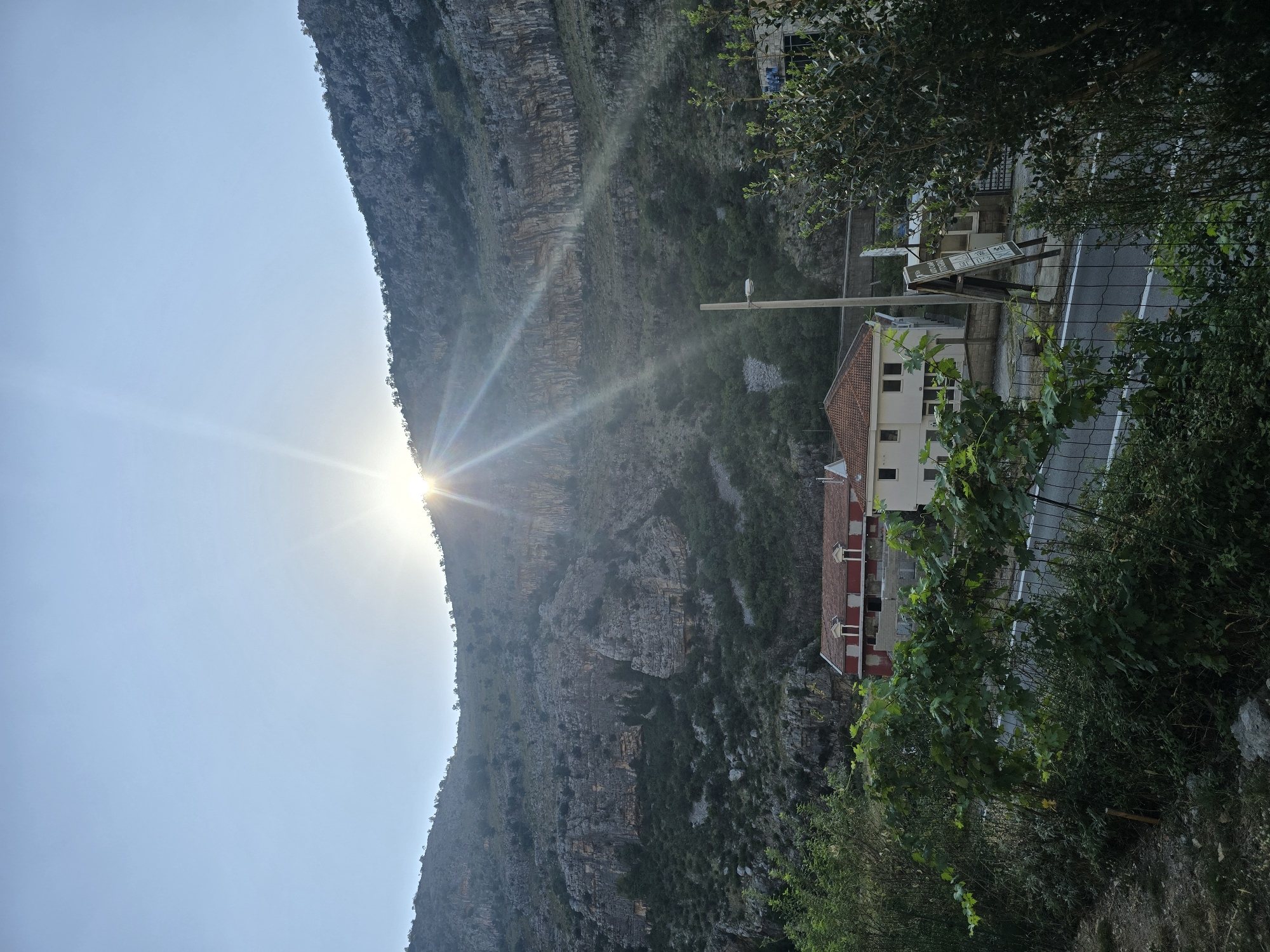 This is a vertical, eye-level, outdoor shot of a mountain valley in Montenegro, likely near Podgorica, bathed in the bright glare of the sun emerging from behind a rocky mountain peak. The sky is a pale blue, with the sun creating a strong lens flare that casts visible light rays downwards. The mountain itself is a dominant feature, its sheer rock face etched with horizontal layers and covered in sparse vegetation and trees. Below the mountain, several buildings are visible, including a prominent red-roofed building with white trim and multiple windows, and a lighter-colored building to its right. In the foreground, lush green foliage, including a vine-covered fence, obscures a paved road with white markings. A lamppost stands between the buildings and the mountain.
