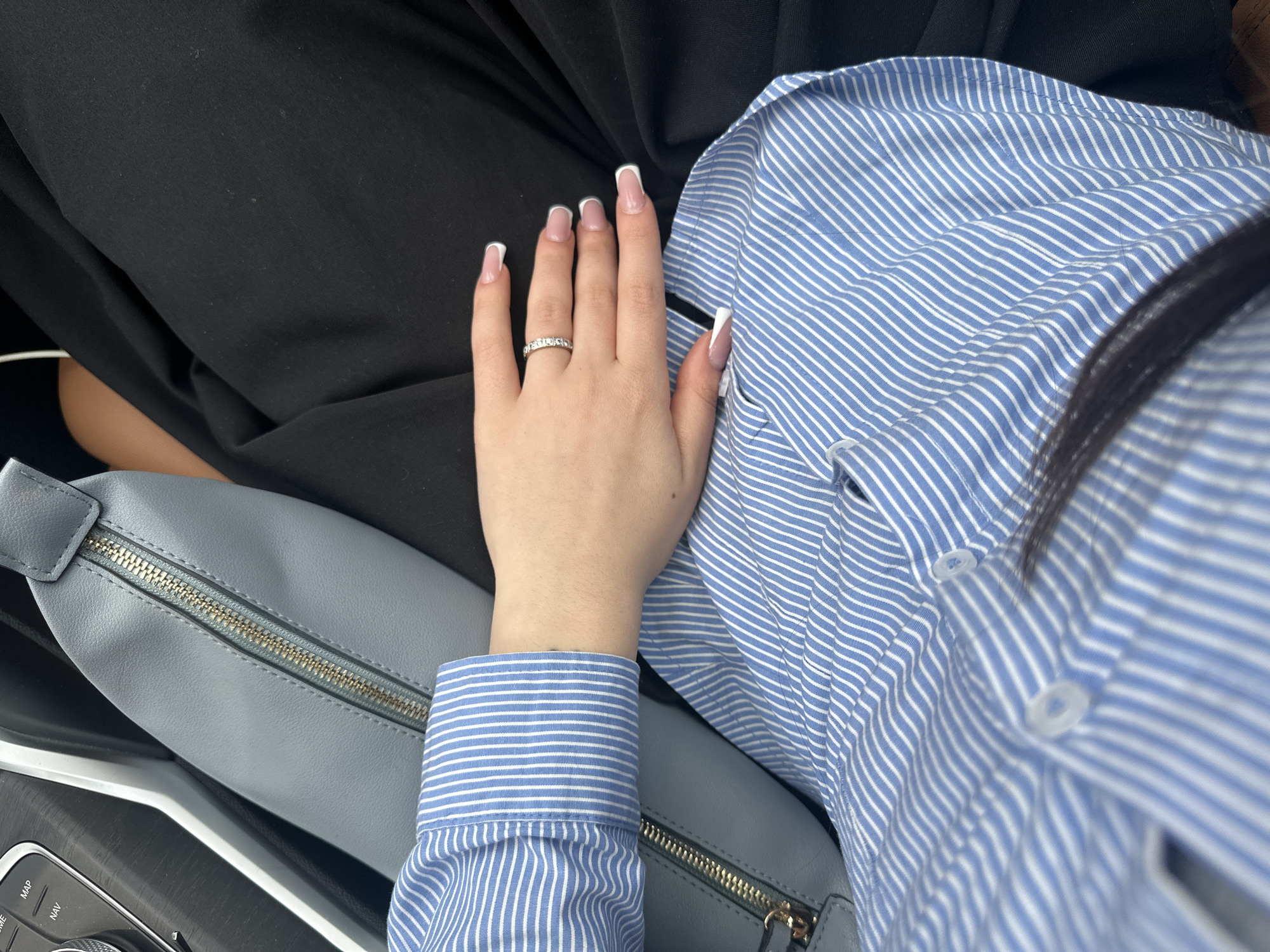 A close-up shot shows a light-skinned hand with French-manicured nails resting on a light blue and white striped button-down shirt. The hand wears a silver ring with multiple stones. The shirt appears to be worn by the person whose hand is visible. To the left of the shirt is a light gray leather-like bag with a gold zipper. Adjacent to the bag is part of a car's interior, featuring a black dashboard with a circular dial and buttons labeled "MAP" and "NAV." The background includes a person wearing black pants or a skirt. The image does not provide enough information to determine the location in Yerevan, Armenia.