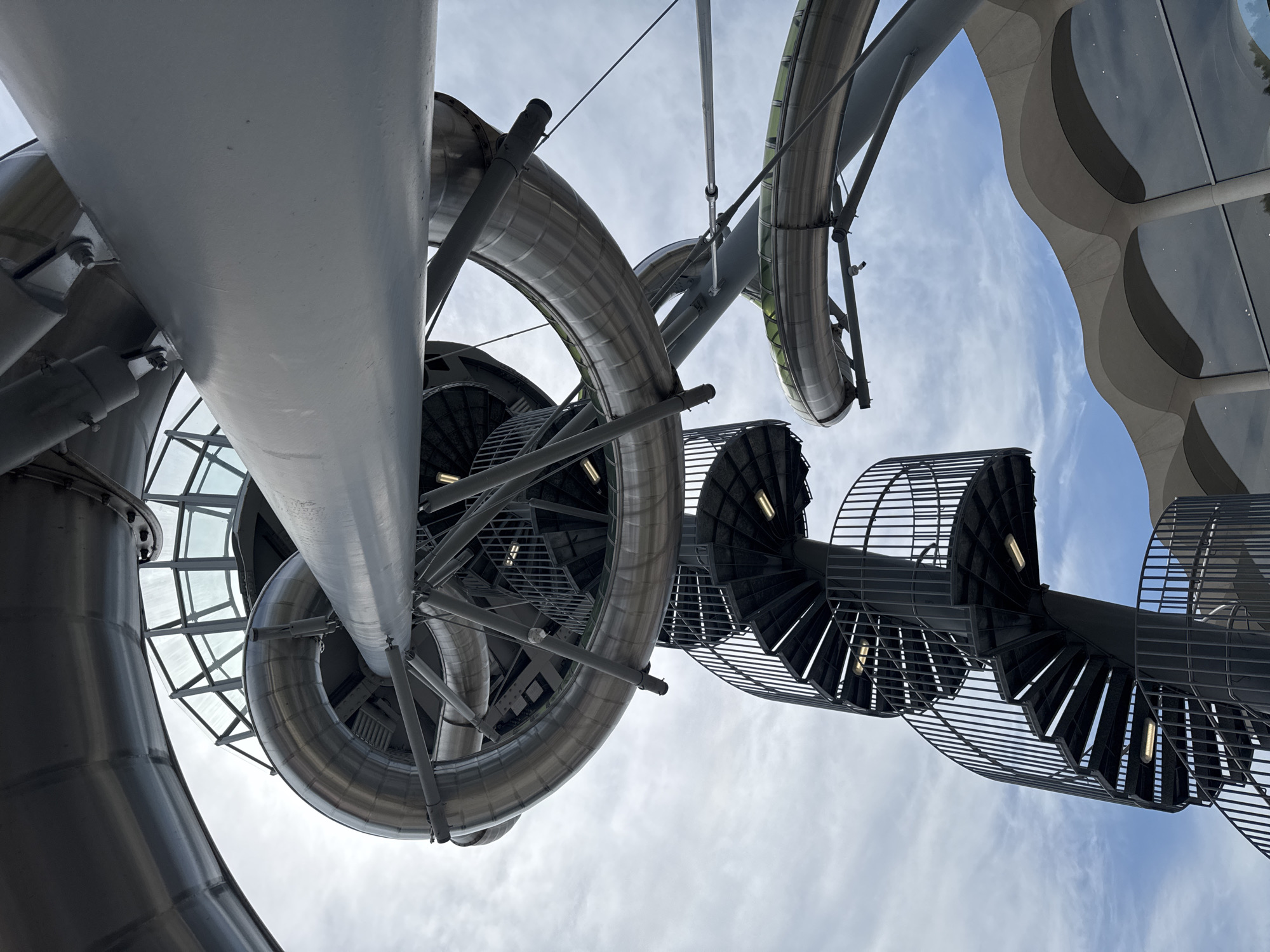 The image shows a low-angle view of a modern architectural structure featuring a winding metal slide and a spiral staircase against a cloudy sky. The slide is made of polished metal and appears to be enclosed in some sections with clear panels. The spiral staircase is dark grey metal with railings and lights visible on the steps. The structure is complex, with multiple levels and curves, suggesting it might be a public attraction or part of a building with recreational facilities. The sky is mostly covered with white and grey clouds. Below the metal structure, there is a glimpse of a building with a distinctive wavy roofline and large windows reflecting the sky.
The location is identified as the city of Aventura, United States of America, which suggests this structure could be a well-known landmark or attraction within the city. Without further information, it's not possible to pinpoint the exact landmark within Aventura.