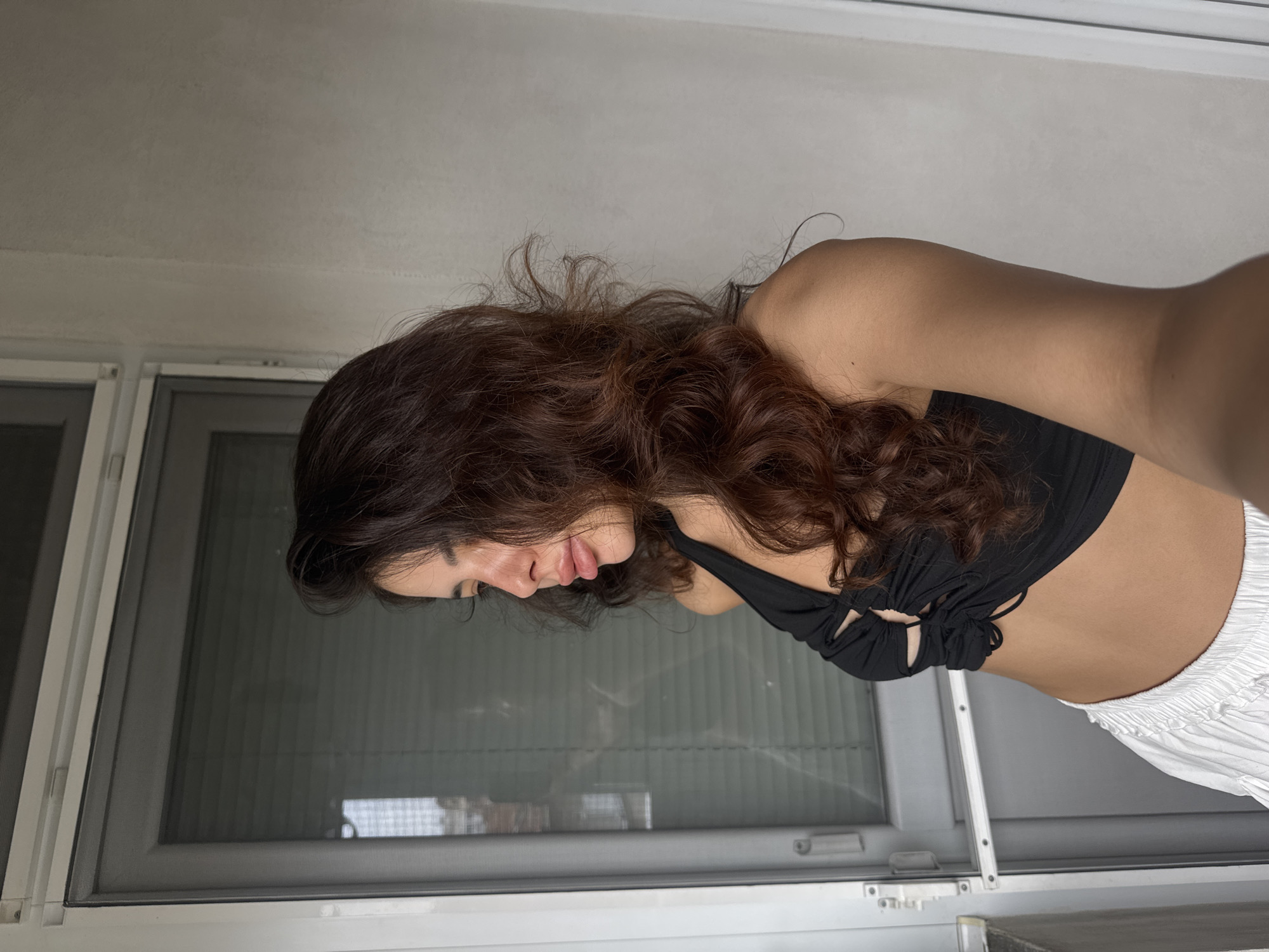 The media file shows a young woman with long, wavy brown hair, leaning forward with her head down, looking towards the right side of the frame. She is wearing a black bikini top and white pants. Her skin is tan, and her hair is partially obscuring her face. The background features a window with blinds, suggesting an indoor setting. The lighting appears to be artificial, possibly from an overhead light source, creating soft shadows. The scene is captured from a slightly high angle, looking down at the woman. There are no visible text elements in the image. The location context provided is Plovdiv, Bulgaria, but there are no specific visual cues within the image to confirm this. The overall mood is subdued, possibly hinting at introspection or relaxation.
