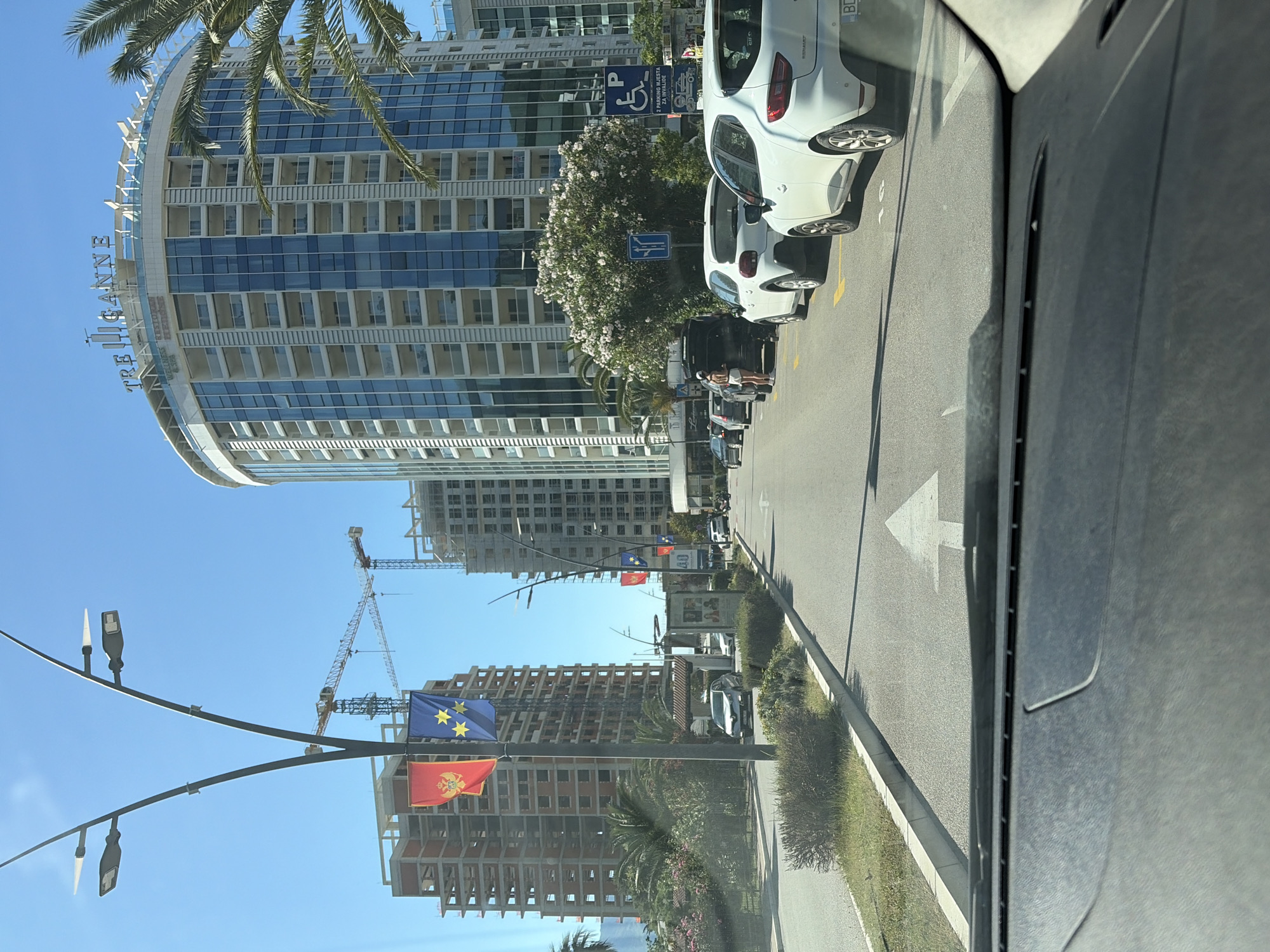 The image shows a street scene in Budva, Montenegro. The view is taken from inside a vehicle, looking through the windshield. On the left side of the street, a large, curved, modern building labeled "TRE CANNE" is visible. Further down the street are several buildings under construction, with cranes present. The flags of the European Union and Montenegro are displayed on a lamppost. Cars are parked along the right side of the street, and a few people can be seen in the distance. The street has an arrow marking indicating the direction of traffic. Vegetation lines the right side of the road. The weather is sunny and clear.