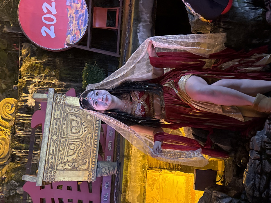 A female subject is depicted in an elaborate costume on a rocky platform, likely a performance stage, in an outdoor or semi-outdoor setting at night, within Yuzhong District, China. The subject, exhibiting white theatrical facial makeup and dark braided hair adorned with a silver-colored headpiece featuring a central red gem, wears a red and gold two-piece ensemble. This attire consists of an embroidered bustier and a flowing, layered skirt, which is split to reveal her right thigh. A sheer, light-colored veil is draped over her head and shoulders, held by her hands, and she wears light brown ankle boots.
The background elements are artificially illuminated. To the subject's left stands a large, golden, ornate structure resembling an ancient Chinese ritual vessel, positioned in front of a red panel with visible Chinese characters. Behind this, a yellow-lit stone wall is visible. To the subject's right, a large red circular object, designed like a drum, displays "2020" in white digits and a panoramic cityscape illustration; it is supported by a dark frame. The upper background features artificial waterfalls cascading over dark, rock-like formations amidst green foliage, all brightly lit with warm yellow and orange lights. The immediate foreground consists of dark, textured rock formations. A black box, possibly a speaker or stage light, is situated on the ground to the left of the subject. The subject maintains a neutral expression, looking slightly to her left (viewer's right).