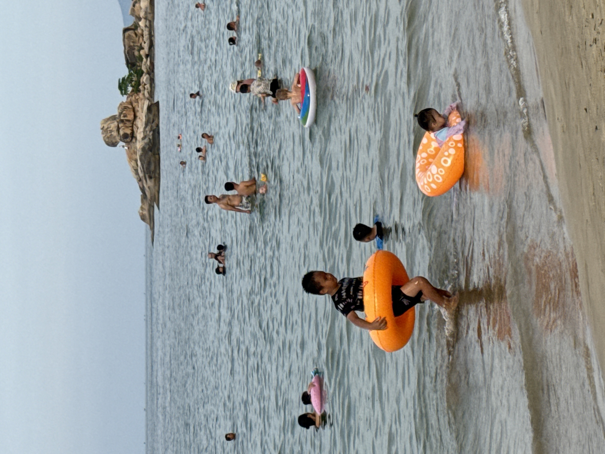 In the ocean off the coast of Nha Trang, Vietnam, several people are enjoying the water. A young boy in the foreground is walking in the shallows with an orange inflatable ring. Further out in the water, other individuals are swimming, some with inflatable rings. In the background, a rocky island with a small tree is visible. The sky is overcast, and the water is calm.