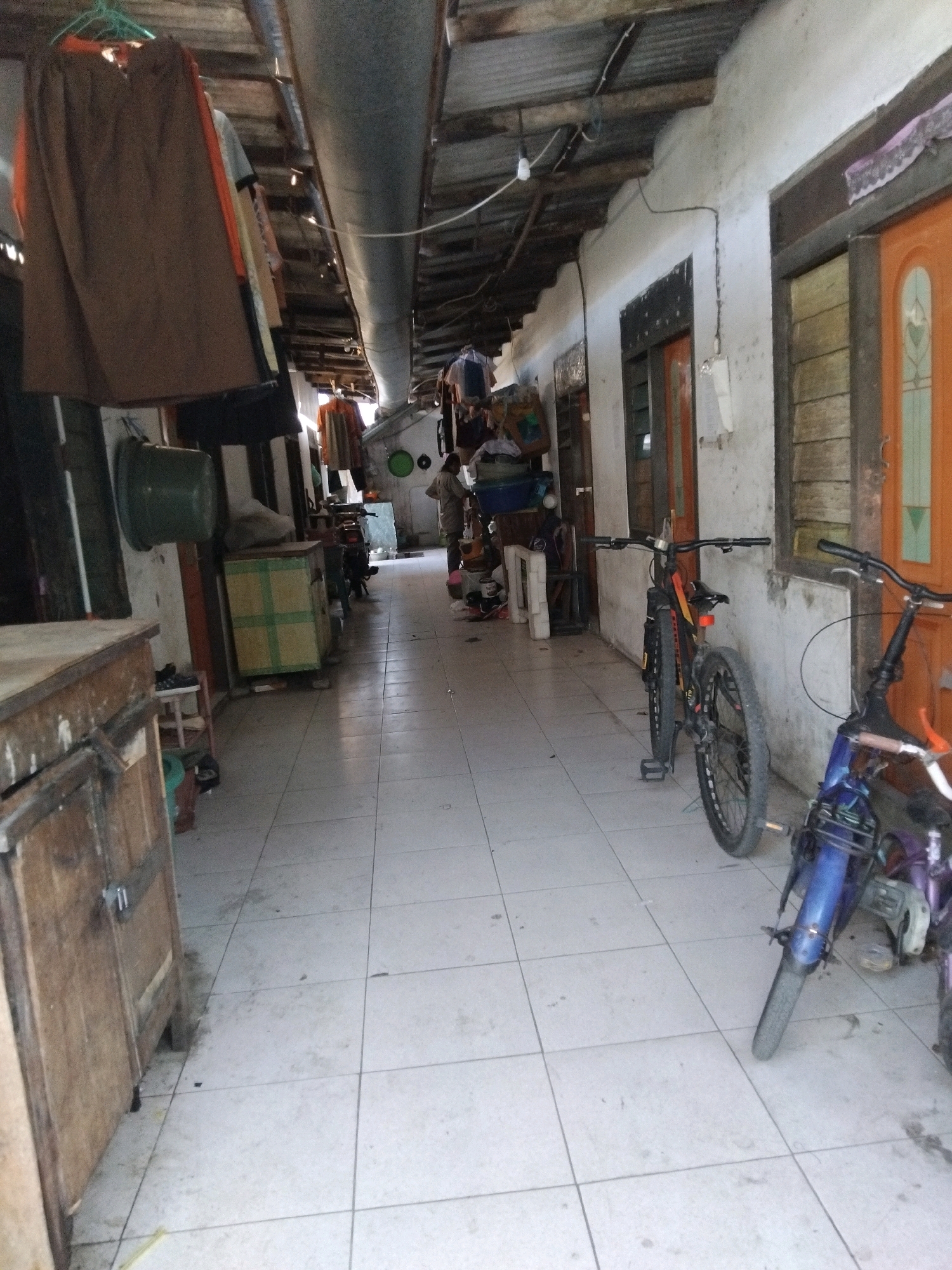 The image depicts an outdoor hallway in a residential area, likely in Berbek, Indonesia, with a tiled floor and a corrugated metal roof supported by wooden beams. On the left, a wooden cabinet is visible, and further down the hall, several items are hanging from a clothesline, including a brown skirt. Various objects are placed along the hallway walls, such as a green plastic tub, a large cardboard box with green stripes, and what appears to be a motorcycle parked further down. Two bicycles are positioned near the right wall; one is black and orange, and the other is blue and purple. A person is standing in the middle of the hallway, near the motorcycles, appearing to be interacting with them or passing by. Doors are visible on the right side of the hallway, one of which is orange. The environment appears somewhat cluttered and utilitarian, suggestive of a modest living space or a shared communal area.