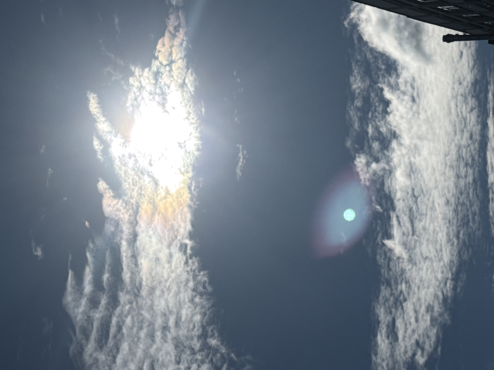 This is an outdoor, daytime photograph looking up at a cloudy sky. The sun is partially obscured by clouds in the upper portion of the image, casting bright rays and creating a lens flare. There are scattered clouds throughout the sky, some appearing thin and wispy, others more dense. In the lower right corner, the dark facade of a building with windows and a chimney is visible. There is a bright, circular lens flare with a blue and pinkish hue in the lower middle section of the image. The image appears to be taken in an urban environment.