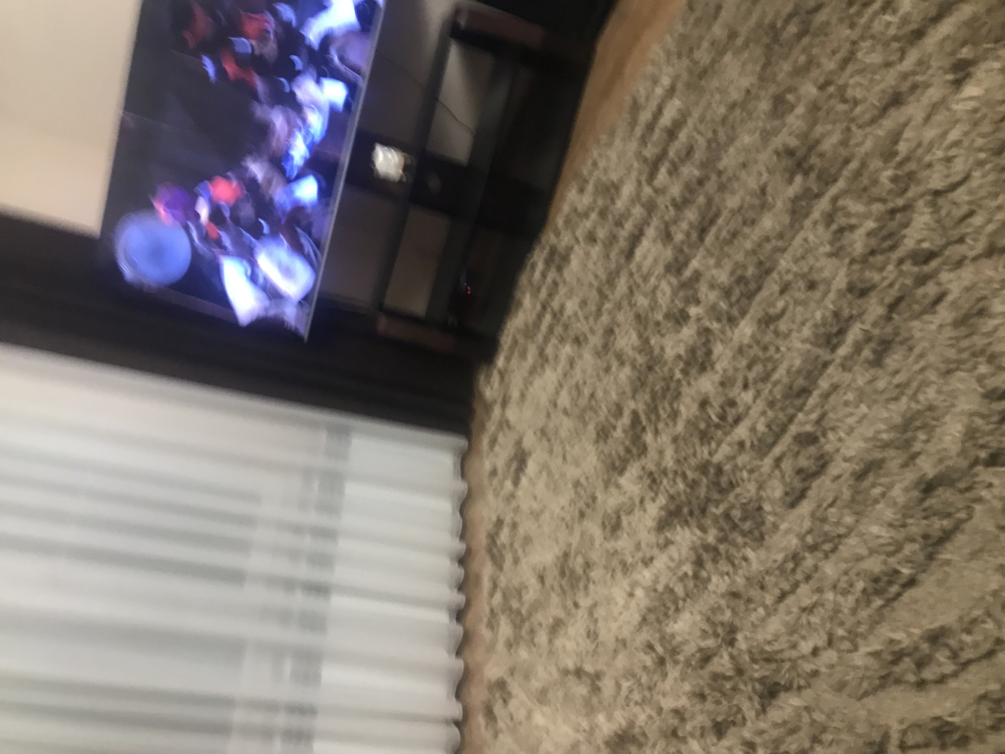 The image shows a room with a carpeted floor, a television displaying an image of people, a dark-colored TV stand, and white window blinds. The location is unknown and no details suggest Yerevan, Armenia.