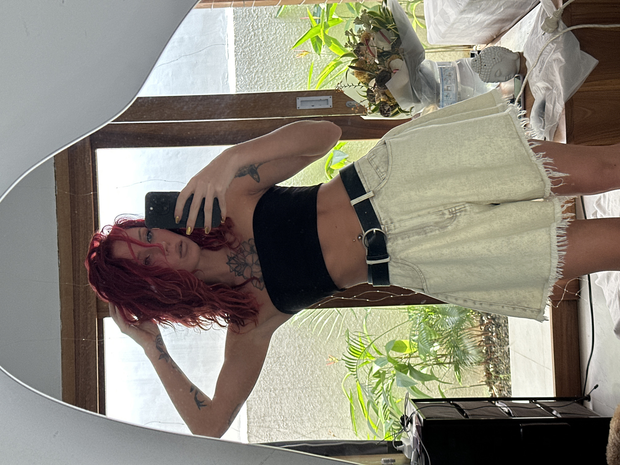 A red-haired woman is taking a selfie in a mirror. She is wearing a black tank top and light-colored denim shorts with a black belt. The woman is indoors, in front of a window with plants visible outside. There is a bouquet of flowers, a Buddha head statue, and other items on a table behind her. The location may be in North Kuta, Indonesia, given the tropical foliage outside the window.