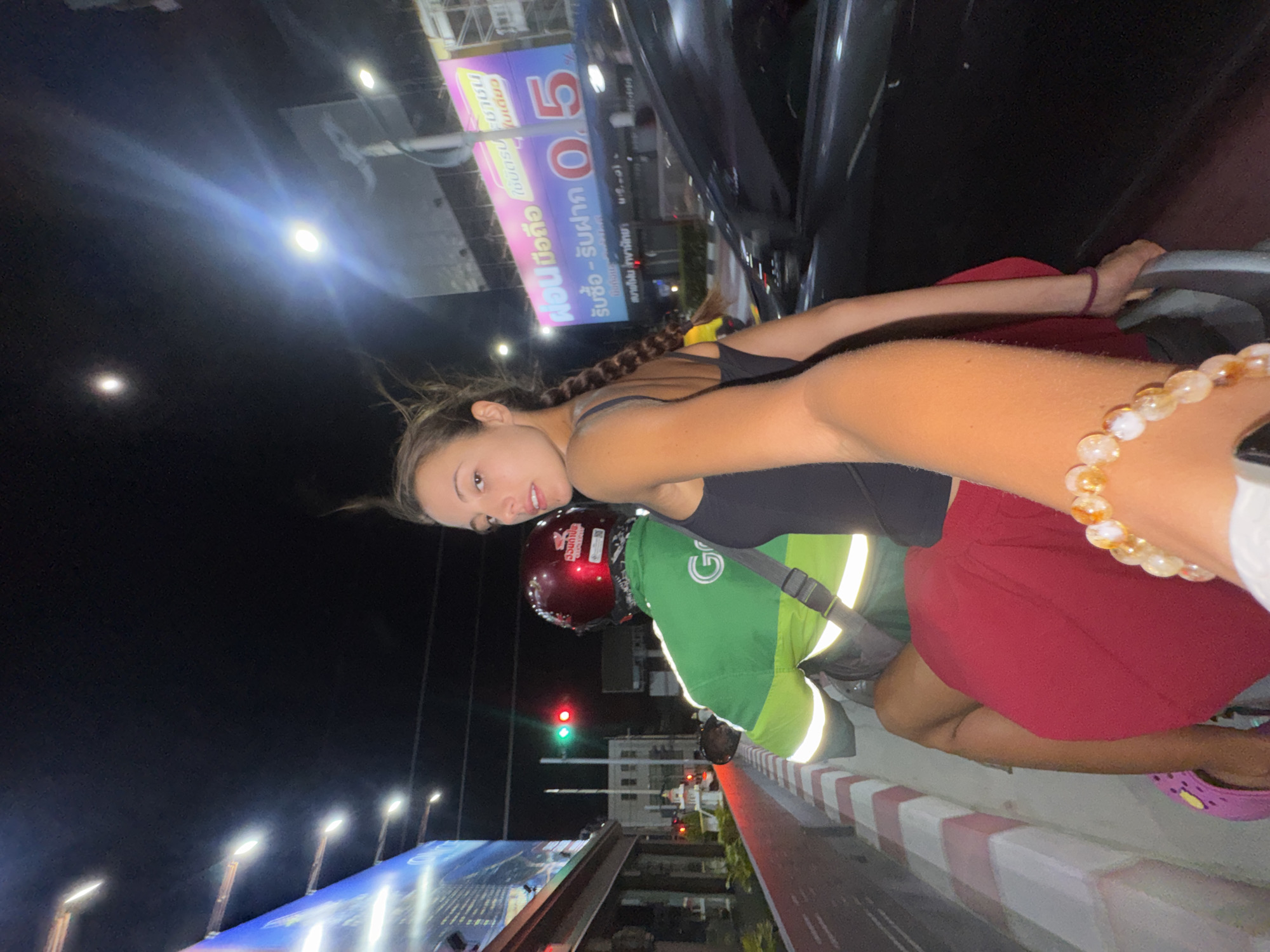 A woman with her hair in a braid is riding on the back of a motorcycle with a male driver in Pattaya City, Thailand. She is wearing a black tank top, a red skirt, and a beaded bracelet. The driver is wearing a green and white uniform and a red helmet. They are stopped at a traffic light on a city street at night. Streetlights illuminate the roadway. There is a large billboard visible in the background. The woman is holding onto the motorcycle and looking at the camera.