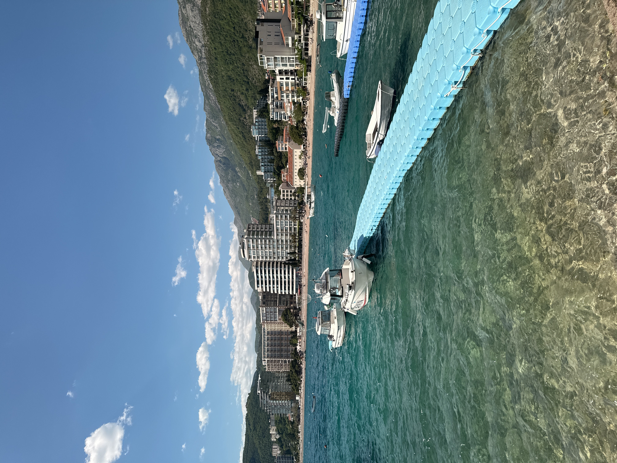 A horizontal shot shows the city of Rafailovići, Montenegro, with a blue pontoon dock extending into the turquoise Adriatic Sea. The scene is bathed in sunlight under a blue sky with scattered white clouds. Along the coastline, various buildings and structures line the shore, backed by a mountain range covered in greenery. A beach is visible between the buildings and the water's edge, with some people visible on the beach. Several white boats are docked along the blue pontoon. The water is clear enough to see some of the seafloor.