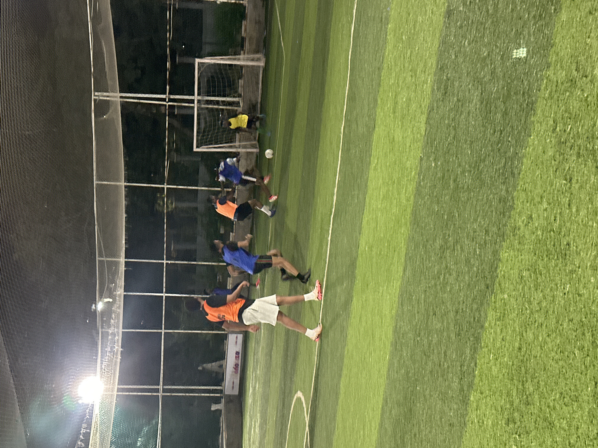 The image depicts an outdoor, artificial turf soccer field at night, illuminated by overhead lights. The field is marked with white lines. In the foreground, a person wearing an orange bib and white shorts is visible, with another person in a blue bib and black shorts running past. Further in the background, two more individuals are playing soccer, with one in a blue bib and the other in a yellow bib. A soccer goal with a net is positioned on the right side of the field. The scene is set within a sports facility, likely a recreational complex in Abuja, Nigeria.