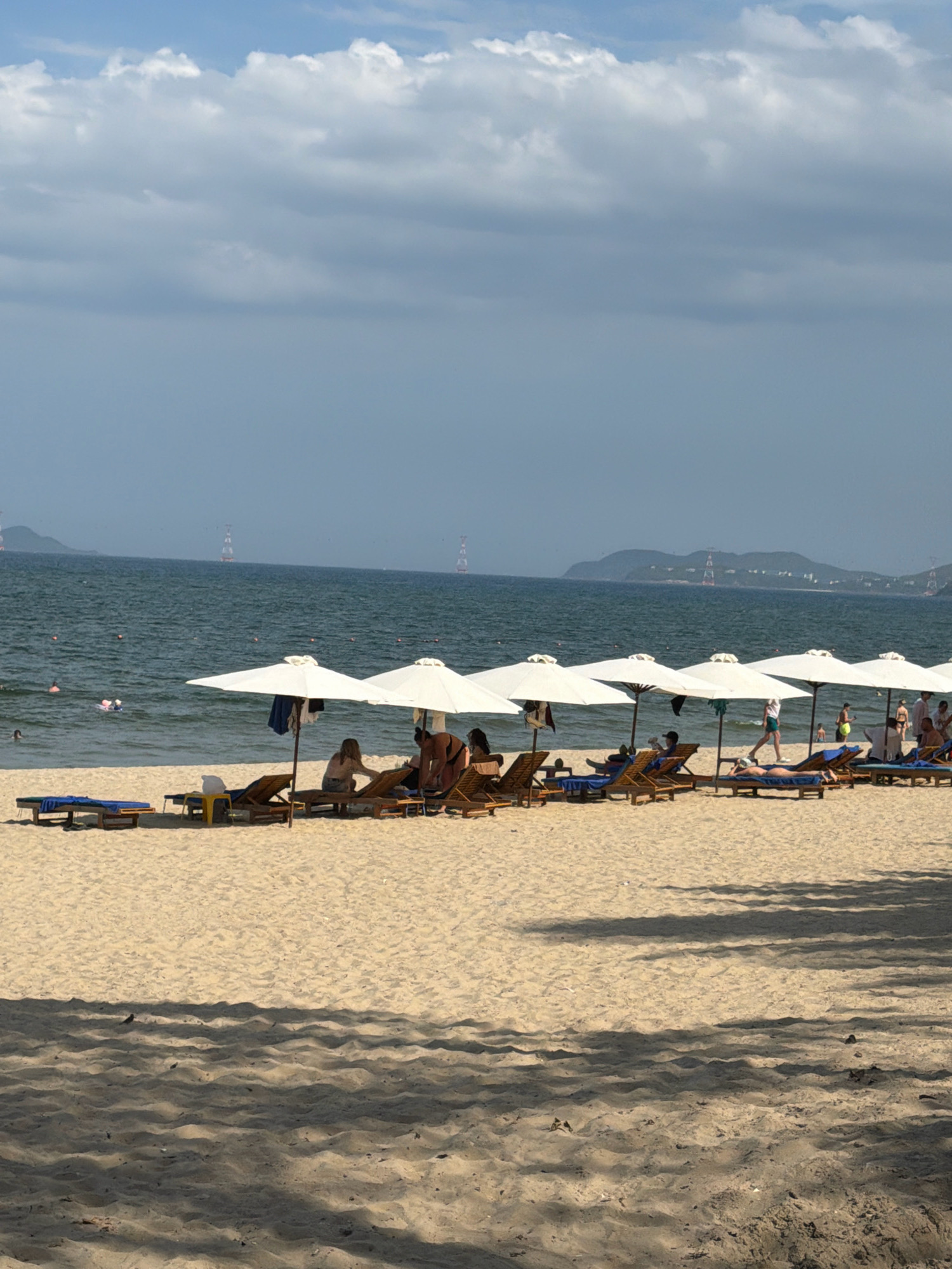 This image depicts a sunny day at a beach in Nha Trang, Vietnam. The foreground is dominated by sand, with a clear shadow of beach umbrellas cast across it. In the midground, a row of white beach umbrellas shades lounge chairs, many of which are occupied by beachgoers. People are seen relaxing on the chairs, with some in swimwear. The ocean stretches out behind the beach, with gentle waves and several people swimming or floating. In the distance, a hazy coastline with rolling hills and tall, industrial-looking towers are visible. The sky is a mix of blue and white clouds. The overall scene conveys a relaxed, vacation atmosphere.