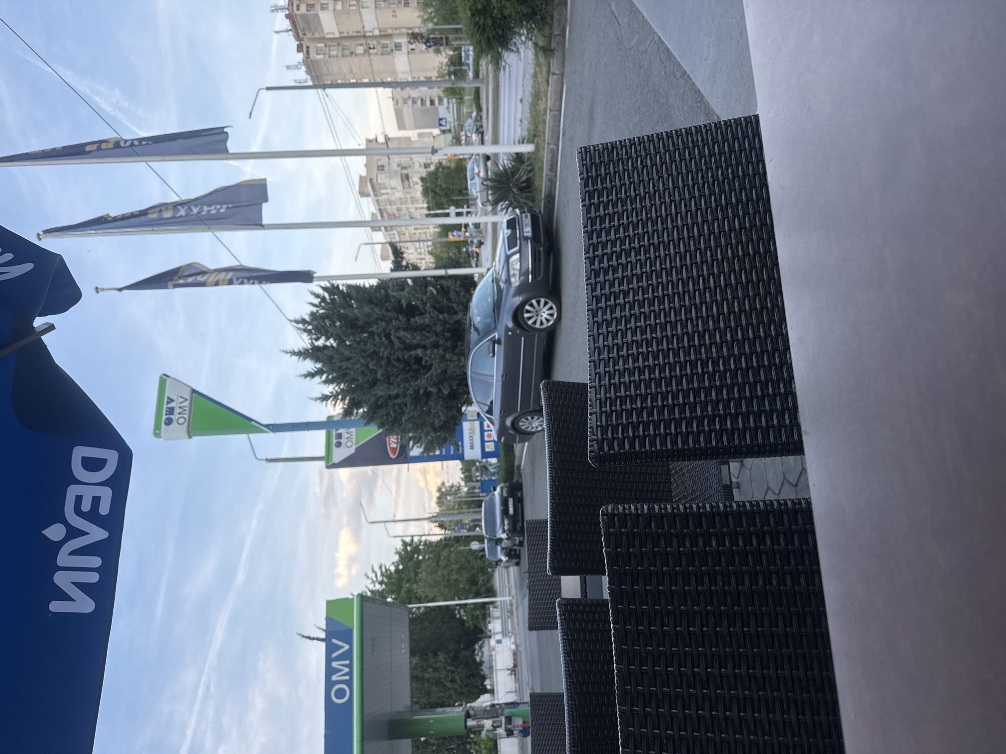 The image shows a view from an outdoor seating area, likely a café or restaurant, in Stara Zagora, Bulgaria. The scene is centered on a row of dark woven chairs, suggestive of an urban street view. To the left, signage for "OMV" and "DEAIN" indicates a nearby gas station. A row of flags is flying from the gas station. Beyond the gas station, a street with parked cars and moving traffic is visible. Buildings line the opposite side of the street, forming the backdrop of the scene. The environment suggests an urban commercial area during daylight.
