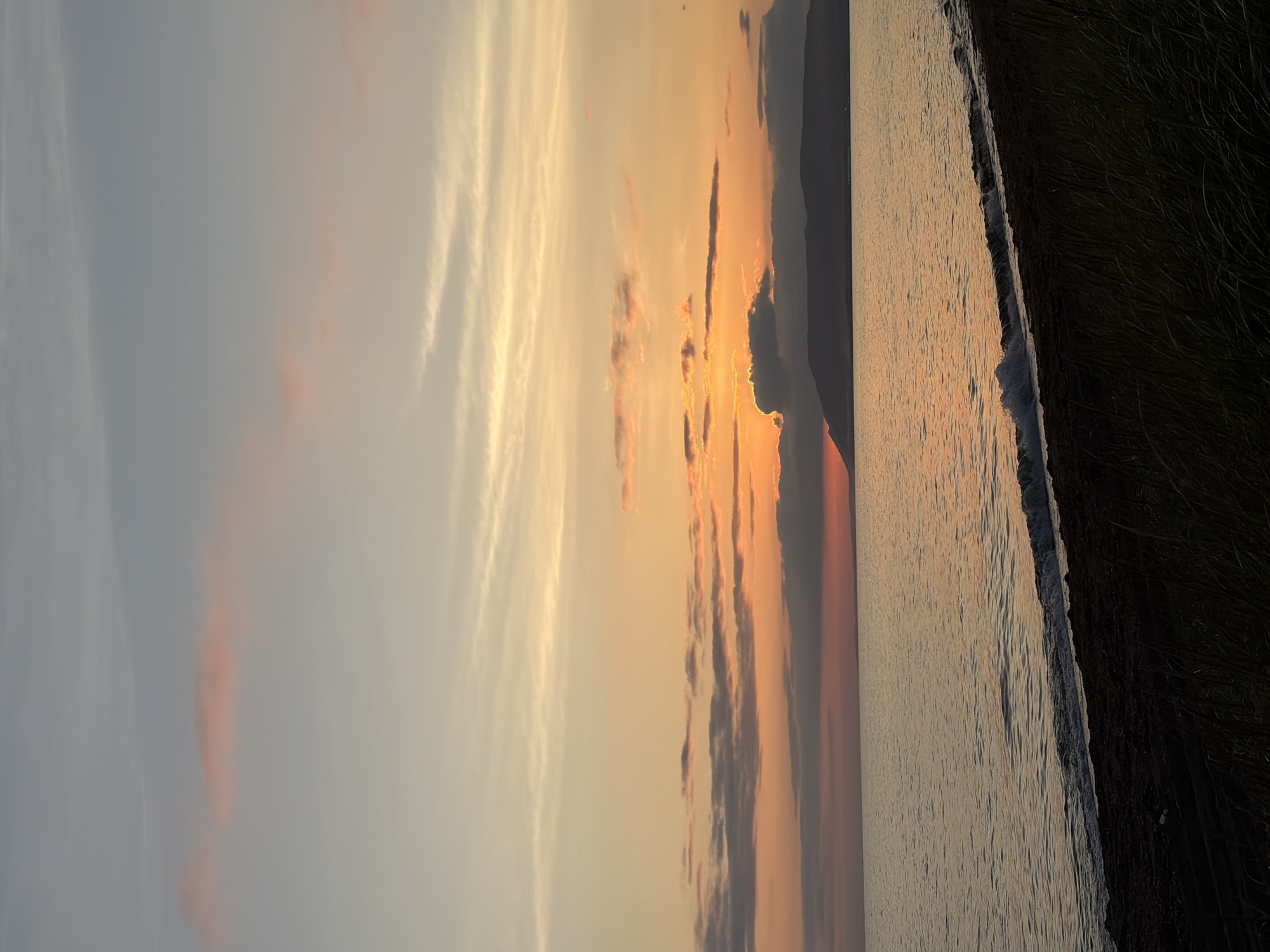 The image depicts a serene sunset over a body of water. The sky is filled with a gradient of colors, ranging from pale blue at the top to vibrant oranges and reds near the horizon. wispy clouds, tinged with pink and orange, are scattered across the sky. A dark, silhouetted landmass, likely a mountain range or hills, is visible in the distance, separating the sky from the water. The water itself is calm, reflecting the warm hues of the sunset. A gentle wave is breaking on the shore in the foreground. The shoreline is dark and appears to be composed of sand or dark soil, with some tall grasses or reeds growing along its edge. The overall impression is one of tranquility and natural beauty. The specific location in the city of Ozerskoe, Russia, is not visually identifiable from the image.