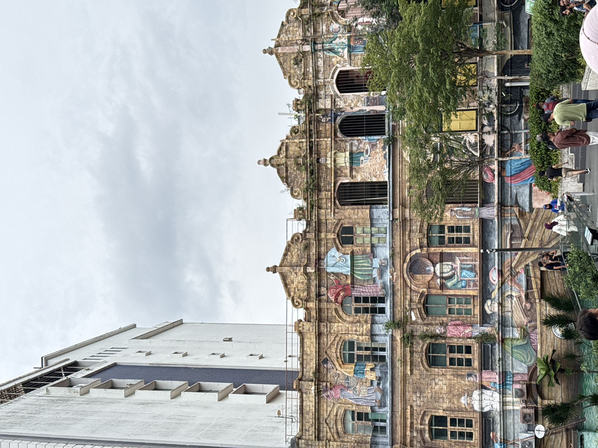The image depicts a view of the Illusion 3D Art Museum in Kuala Lumpur, Malaysia, showing both the decorated facade of the museum building and the adjacent white building under a cloudy sky. The museum facade is designed to create the illusion of a multi-story, old-fashioned building, complete with painted details that resemble architectural features, windows, and murals of people and objects. The adjacent white building has a more modern, stark appearance with rectangular windows and a flat roof. Several people are standing and walking along the sidewalk in front of the museum, and some greenery is present around the base of the museum facade. The photograph appears to be taken from ground level, looking up at the buildings.