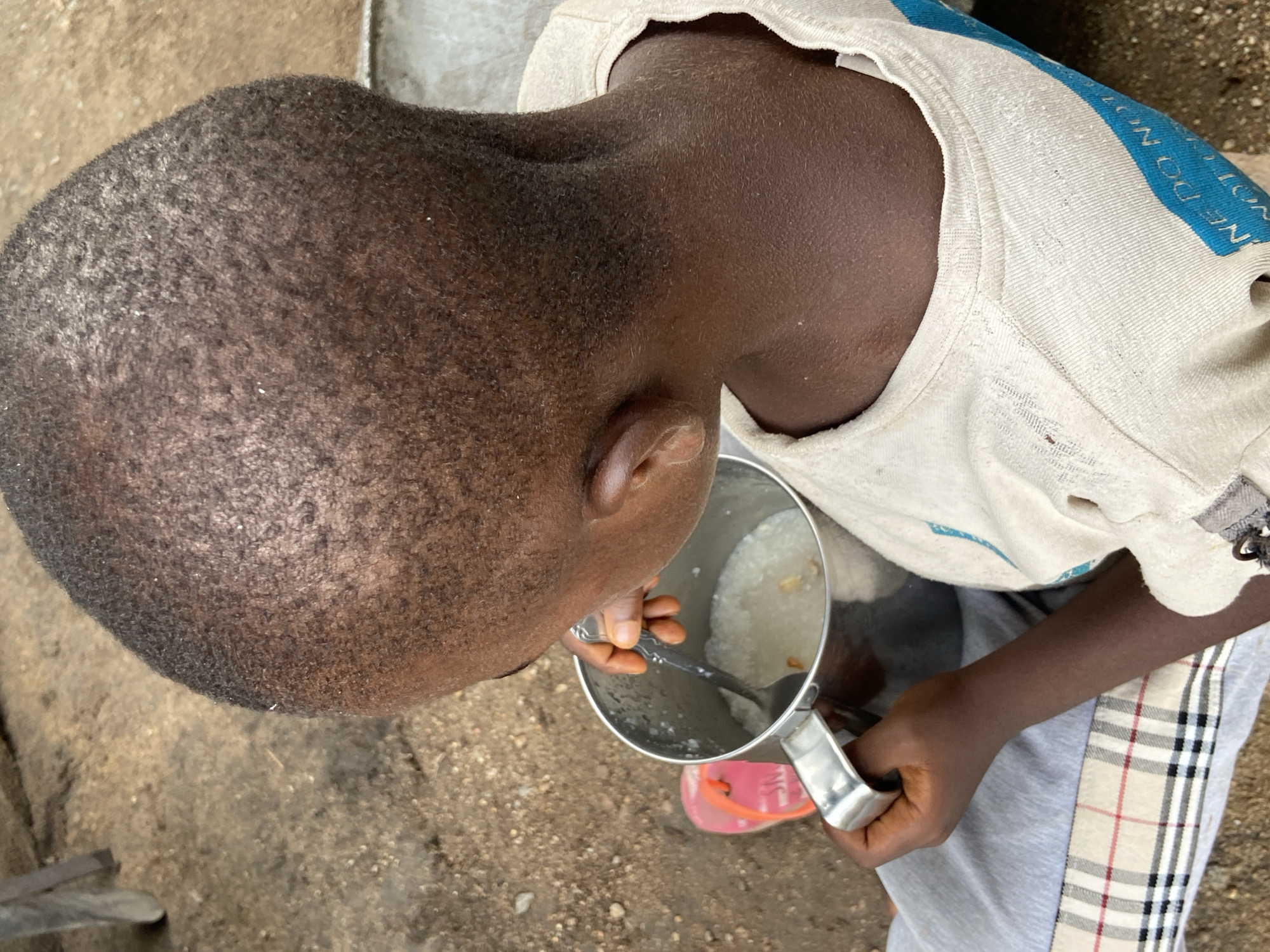 This media file captures a top-down perspective of a young child, likely male, eating outdoors in Mararaba, Nigeria. The child has dark skin and very short, dark hair. They are wearing a light-colored t-shirt, which has a blue section on the right shoulder with partial white text that appears to be "WE DO NOT" (possibly reversed or mirrored). The child also wears grey pants with a distinctive beige, black, and red/pink plaid stripe running down the side.
The primary activity is the child eating from a silver-colored metal pot or mug with a handle. The pot contains a light-colored, thick, porridge-like food, which includes some yellowish fragments. A spoon is inside the pot, held by the child's right hand. Pink sandals are visible on the ground near the pot.
The setting is outdoors, with the child positioned on rough, earthy ground that appears to be dirt or concrete. The lighting suggests it is daytime, with diffused natural light rather than harsh direct sunlight. The scene conveys a sense of everyday life in a modest, possibly rural or low-income environment. The child's posture indicates concentration on the meal.