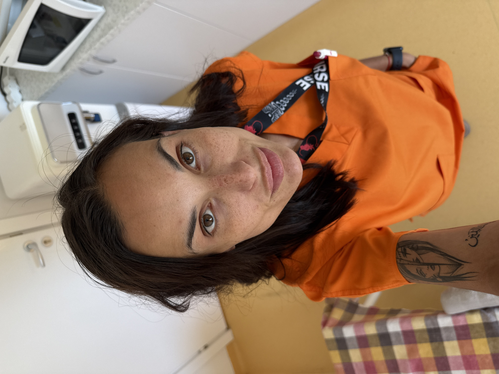 This is a high-angle, eye-level, full-body shot of a woman in an orange scrub top. The woman has dark brown hair, brown eyes, and freckles on her face. She is wearing a black lanyard with red and white text. There is a tattoo on her left forearm. She is standing in what appears to be a medical facility, possibly at a hospital in Pleven, Bulgaria. In the background, a white microwave is visible on a counter, and a water dispenser is to the left of that. A checkered blanket is on a piece of furniture to the left of the frame. The floor is yellow.