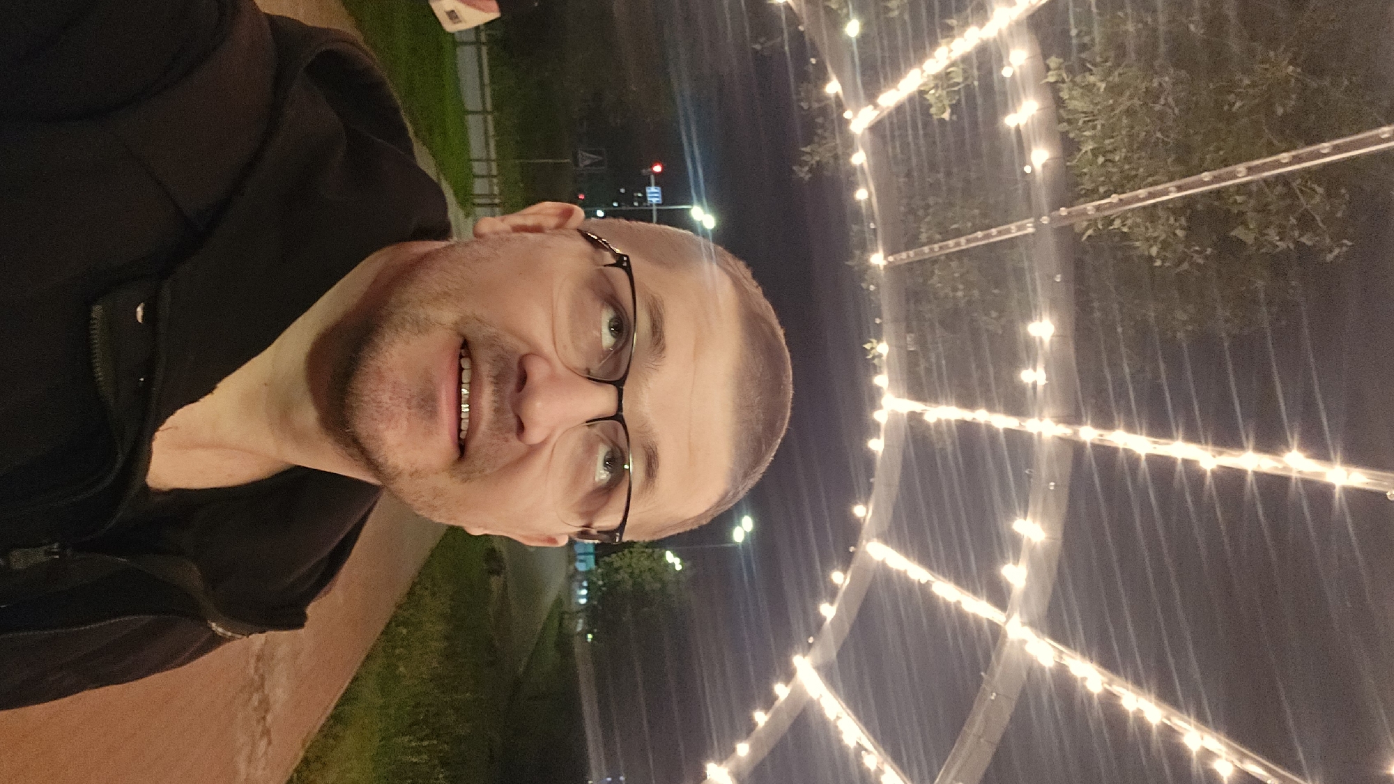 A man in his 40s with short brown hair, a stubble beard, and wearing black-rimmed glasses looks upwards. He is wearing a black hooded jacket over a dark t-shirt. The image appears to be taken at night, possibly on a walkway or street lined with trees. String lights are visible overhead, creating an illuminated archway. In the background, there is a street with traffic lights, a sidewalk, and grassy areas. The overall setting suggests an urban environment. The location is identified as Kyiv, Ukraine, near a pedestrian walkway or park area.