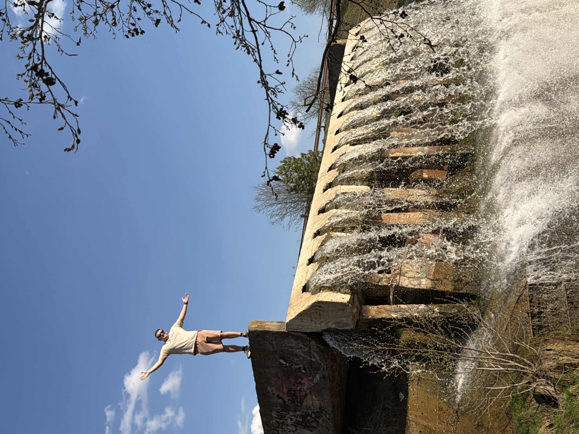 The image depicts a man standing on the edge of a concrete structure at a waterfall in Kamennoye, Russia. The man is light-skinned, wearing a light-colored shirt, brown shorts, and shoes. He is extending his arms outwards. Behind the man is a concrete structure with water cascading over it, forming a waterfall. Branches with leaves are visible in the foreground and background, framing the scene. The sky is blue with some scattered clouds. The image is oriented sideways, with the sky on the left and the waterfall on the right.