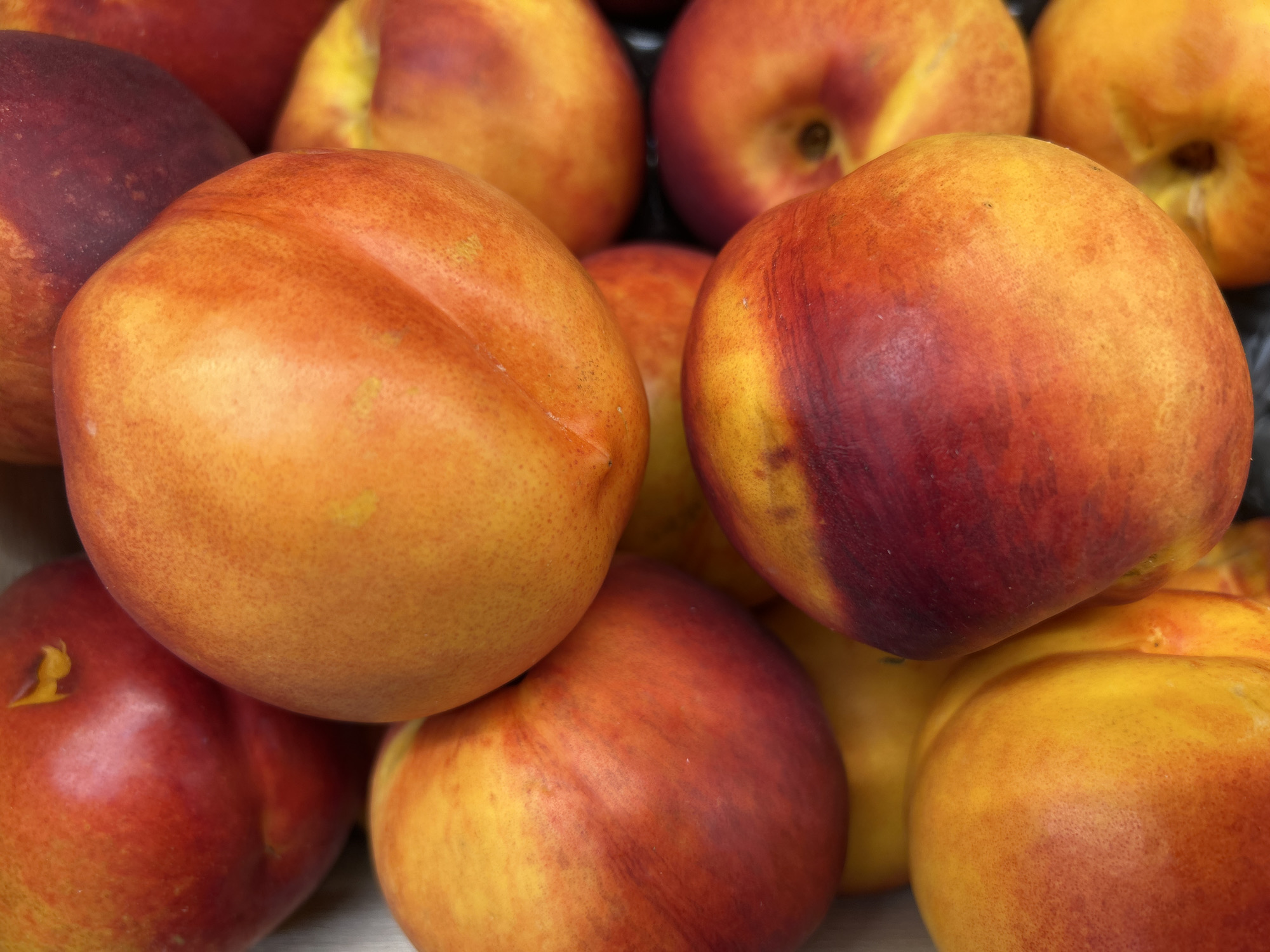 A close-up, overhead view of a pile of ripe nectarines. The nectarines are primarily orange and red, with some yellow hues. Their skins appear smooth and slightly fuzzy. The fruits are stacked on top of each other, filling the frame. The background is dark and out of focus. The lighting is bright, highlighting the vibrant colors of the nectarines. There is no indication of location in Detmold, Germany.