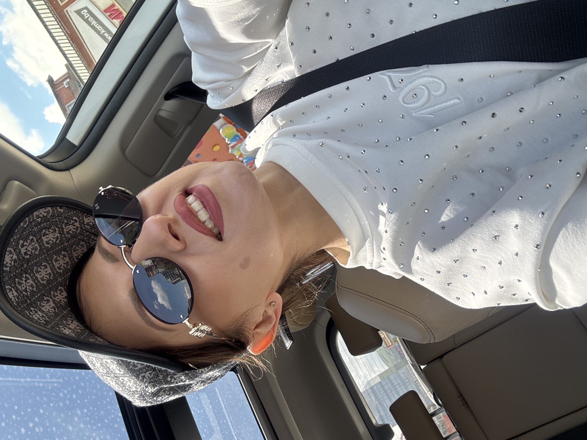 The image depicts a woman wearing sunglasses and a patterned hat, seated in a car, secured by a seatbelt. She is wearing a white sweater with embellishments. Outside the car window, a building with signage is visible, identified as being located in Sofia, Bulgaria. The sky appears partly cloudy.
