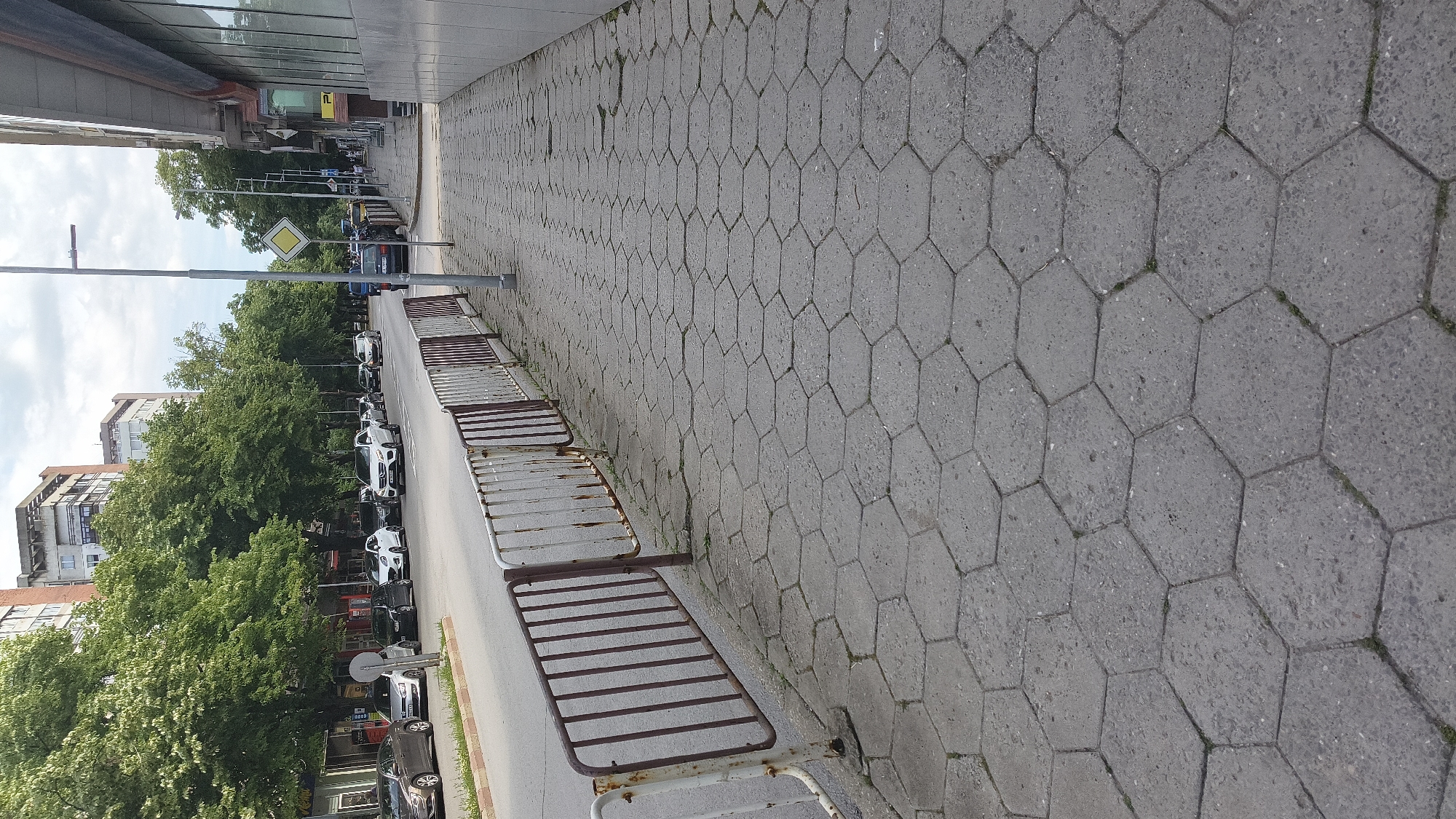 The image depicts a sidewalk paved with hexagonal concrete slabs, bordering a street in Shumen, Bulgaria. A rusty metal barrier separates the sidewalk from the street, where several cars are parked and driving. Mature trees line the street, providing shade. In the background, multi-story buildings are visible. A street lamp and a diamond-shaped yellow traffic sign are present on the sidewalk. The sky is overcast.