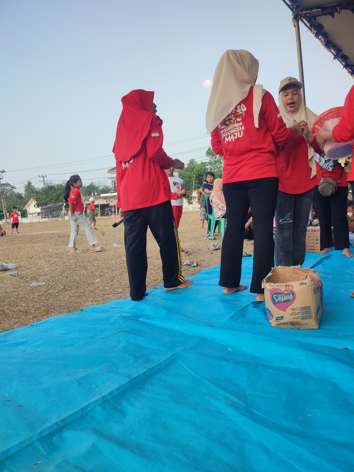 An outdoor, medium-shot, eye-level, full-color photograph captures a scene on the grounds of the city of Nusa Jaya, Indonesia. In the foreground, a bright blue tarp covers the ground. On the tarp, a cardboard box with red and white lettering that reads "Sayang" is open. To the right of the box, a person wearing black pants and a red shirt with "NUSANTARA INDONESIA MAJU" written on the back is visible. To the left of the box, a person wearing black pants and a red shirt is standing. Behind them, a group of people, some wearing red shirts and headscarves, are gathered. A young girl wearing a red shirt and jeans walks across the dry, brown grass in the mid-ground. In the background, there are houses, trees, and power lines, indicating a residential area. The sky is clear and bright blue.